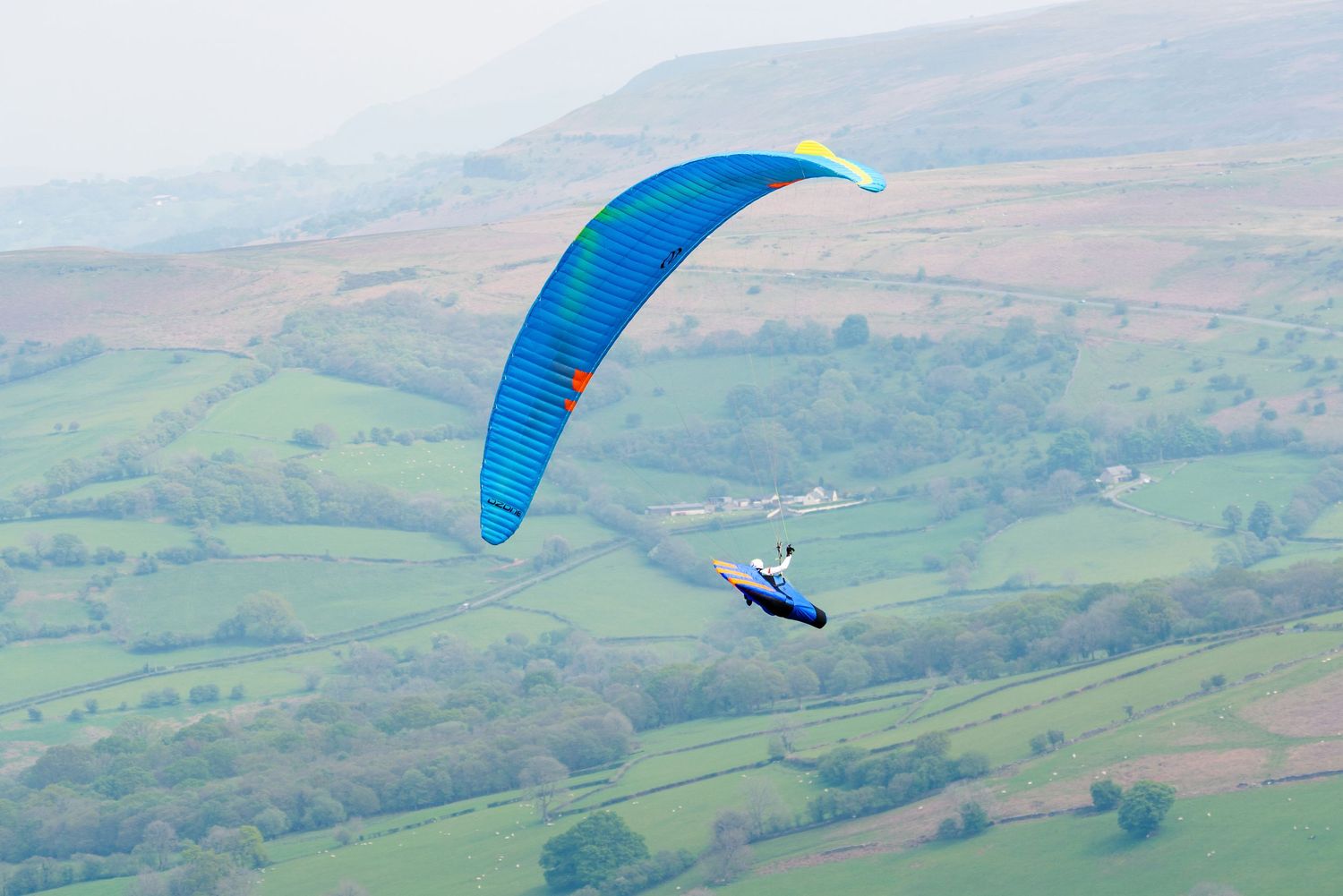 Blue paraglider soaring over pastoral countryside landscape.