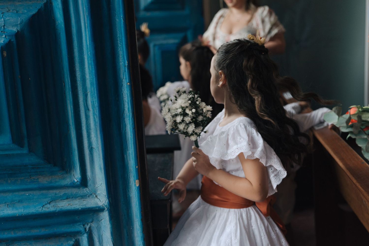 Young flower girls in white dresses with orange sashes waiting at a church wedding.