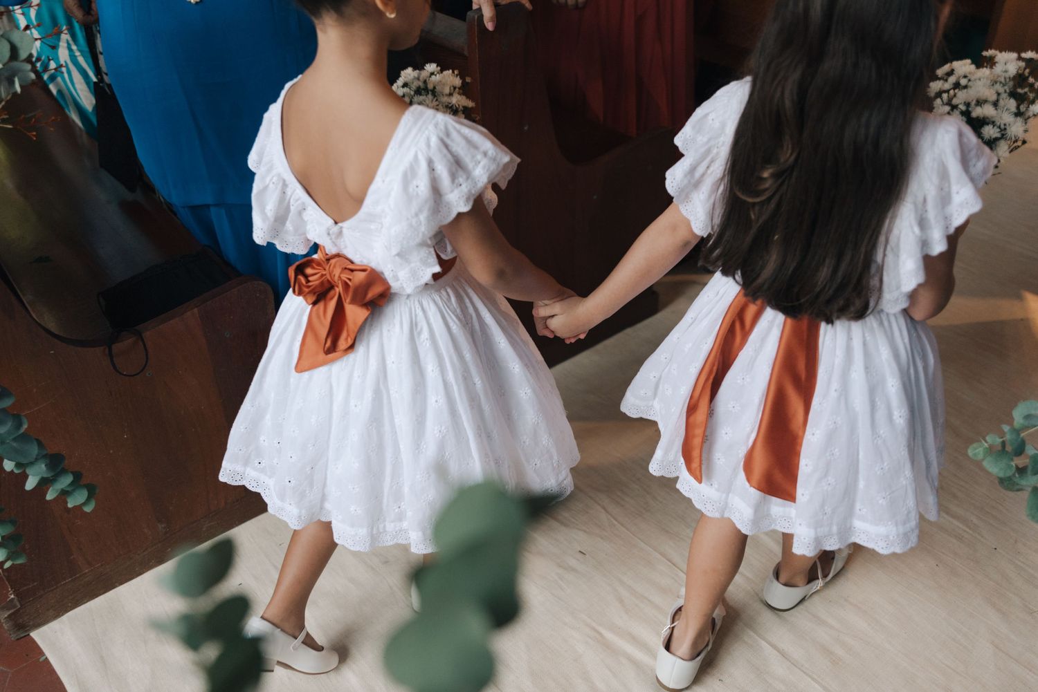 Two flower girls in matching white dresses with orange ribbons seen from behind.