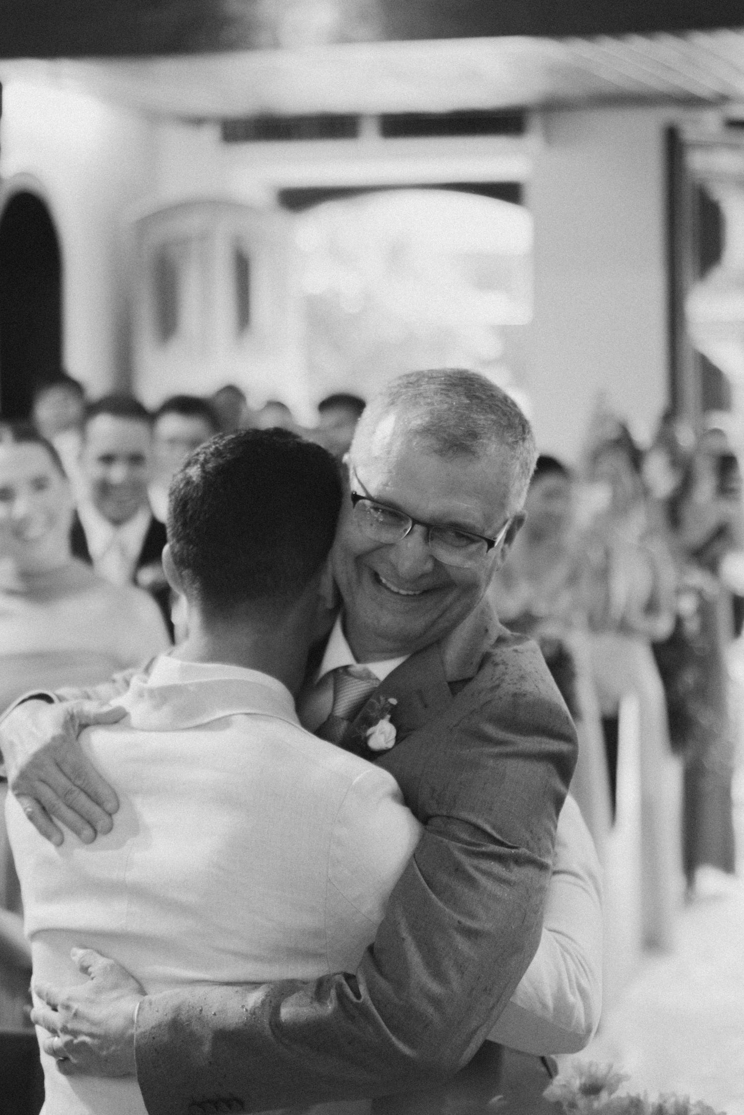 Emotional black and white moment with older man in glasses hugging another person at a wedding celebration.