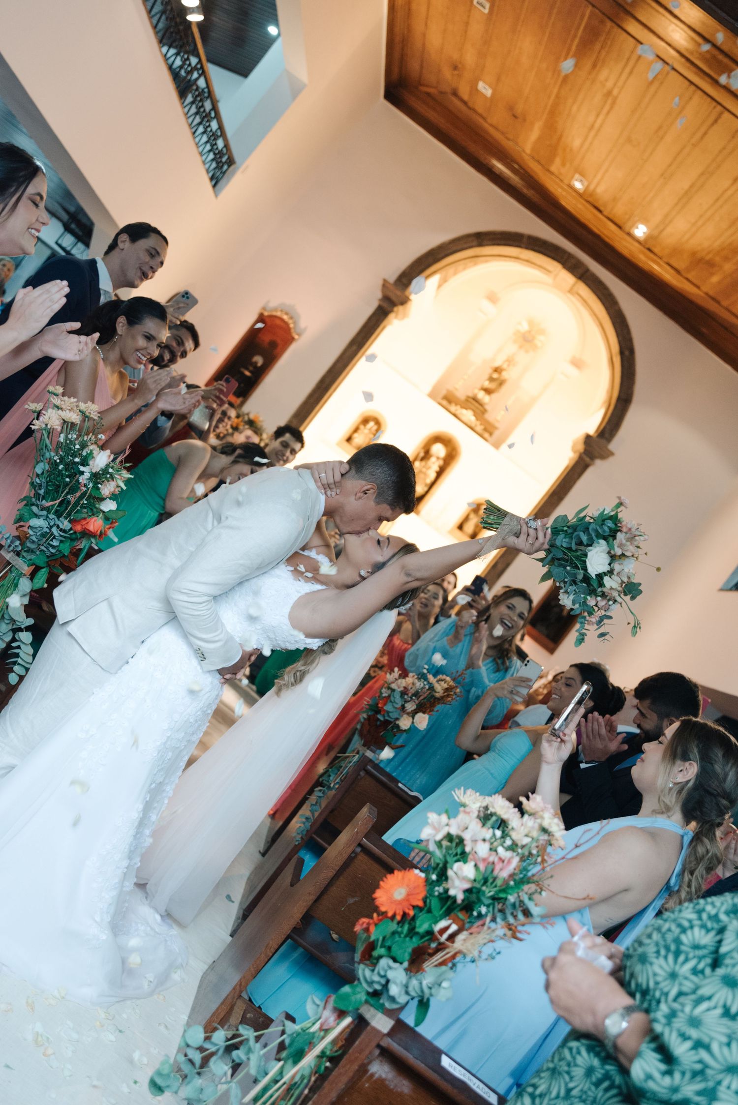 Newlyweds kiss during their wedding ceremony as guests in turquoise dresses watch from the sidelines.