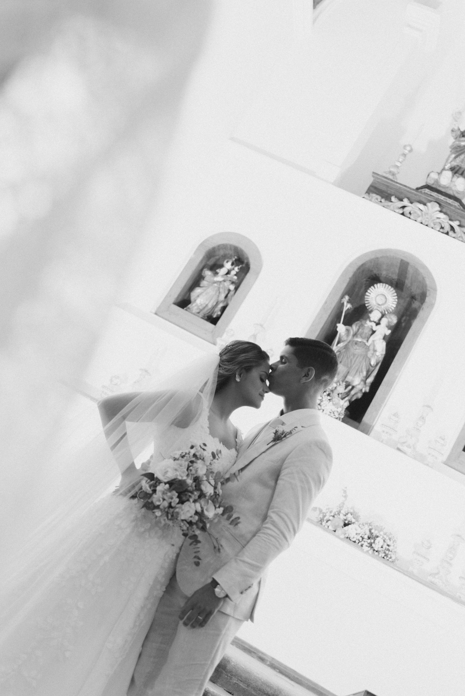 A couple in wedding attire share an intimate moment in a church with religious statues in arched alcoves.