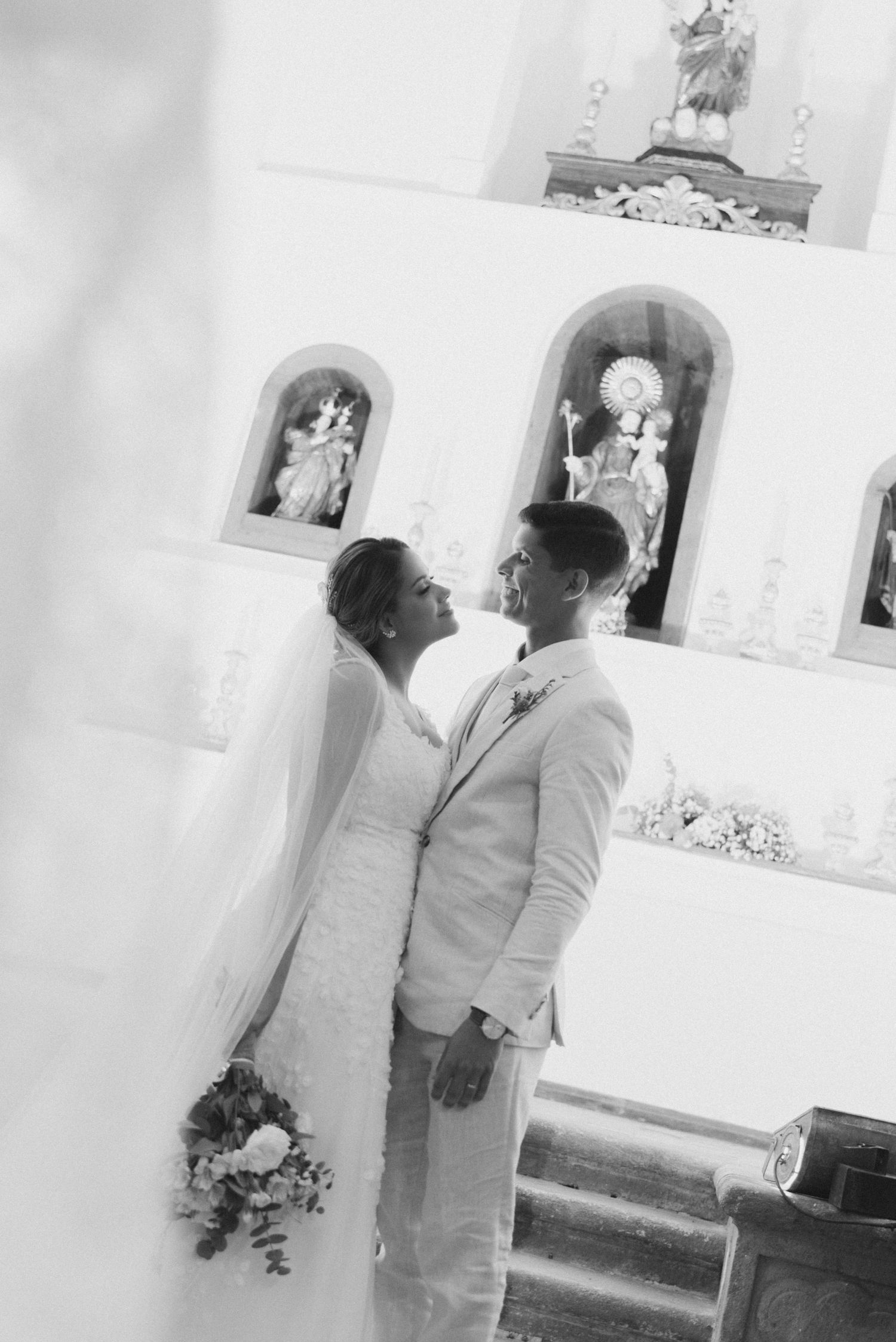 A couple in wedding attire stands together in a church with religious statues visible in the background.