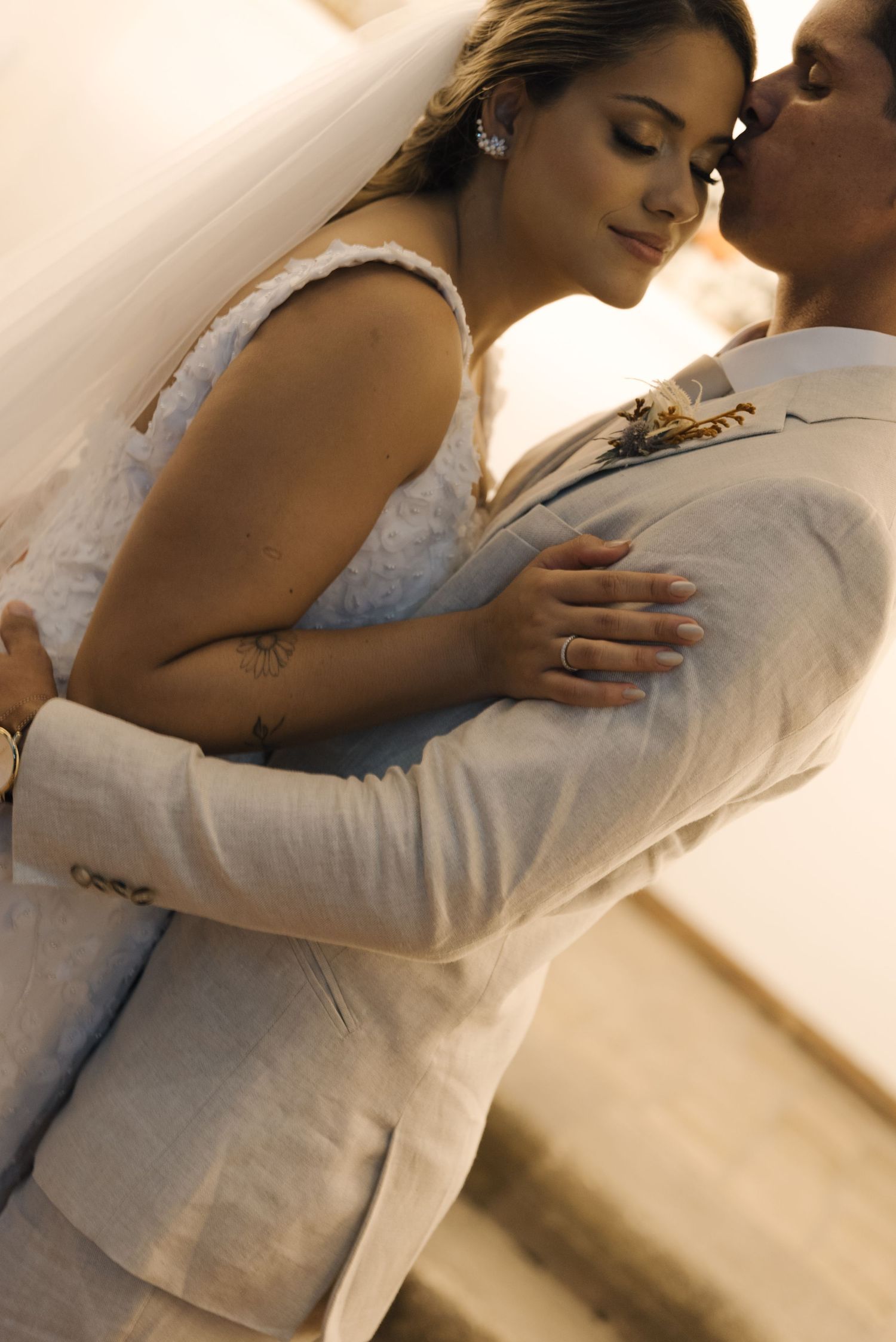 A bride in a lace dress embraces her groom wearing a light colored suit during an intimate moment.