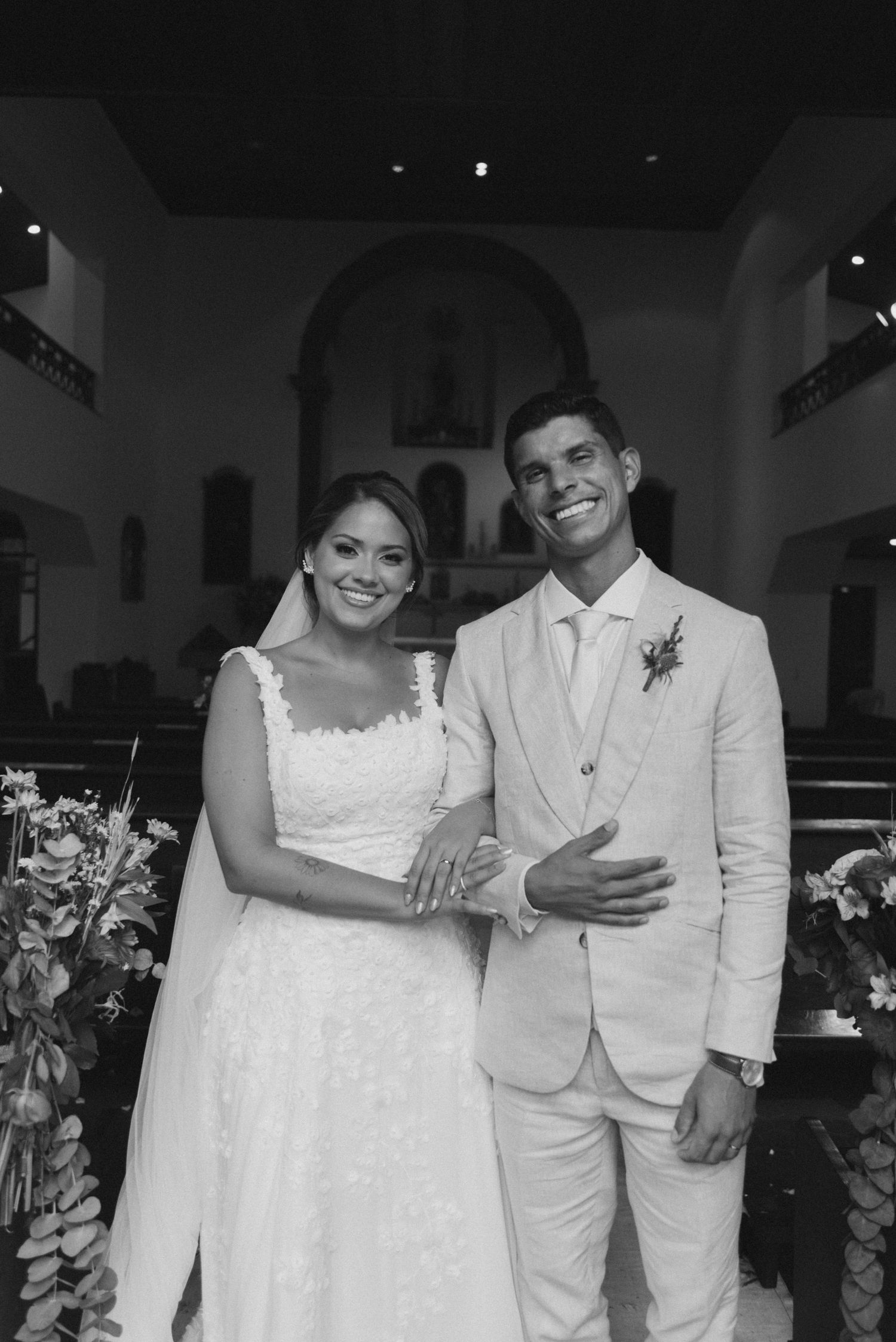 Formal wedding portrait of newlyweds standing together in a church, dressed in white and cream attire.