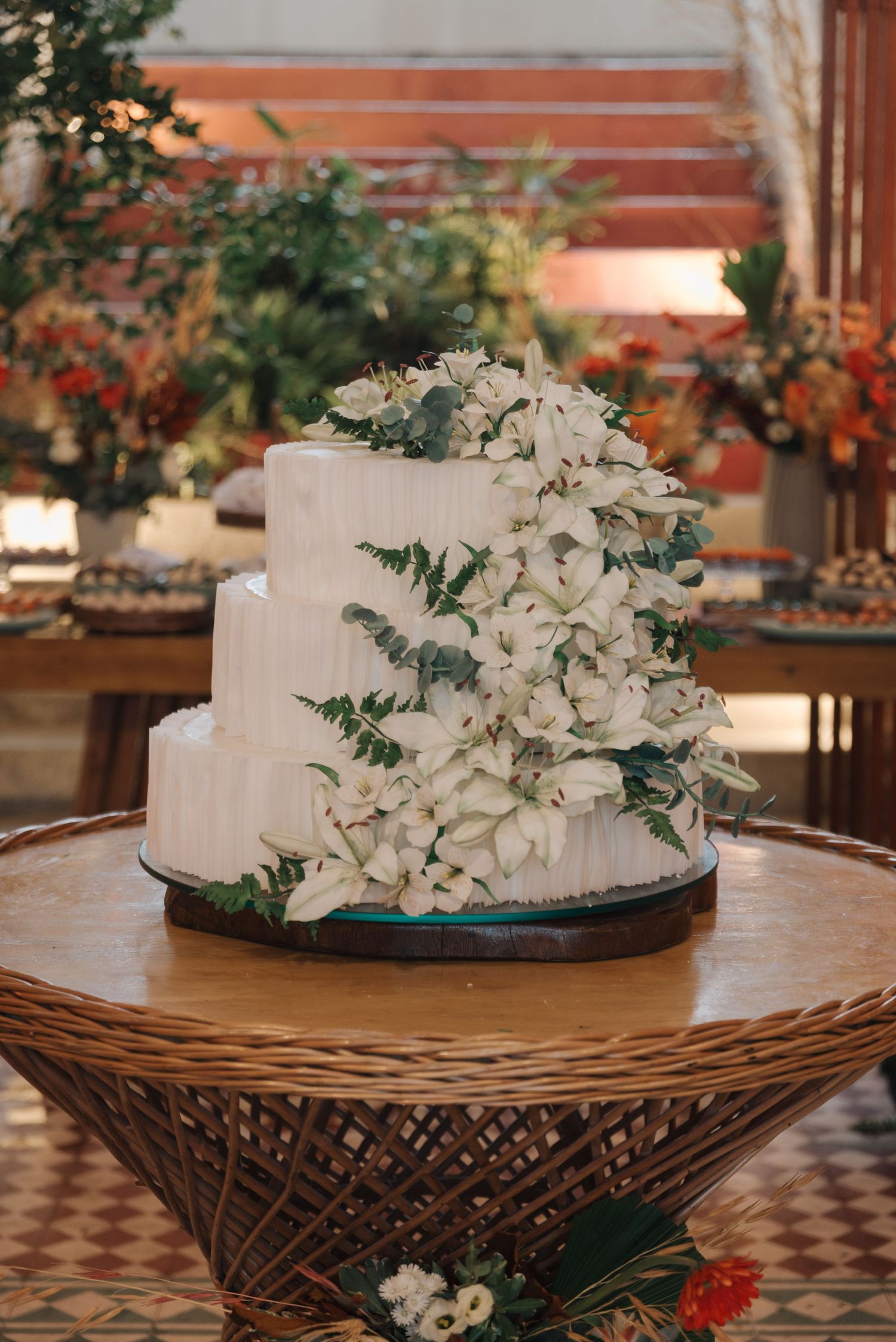 Three-tiered white wedding cake decorated with cascading flowers and greenery on a woven table.