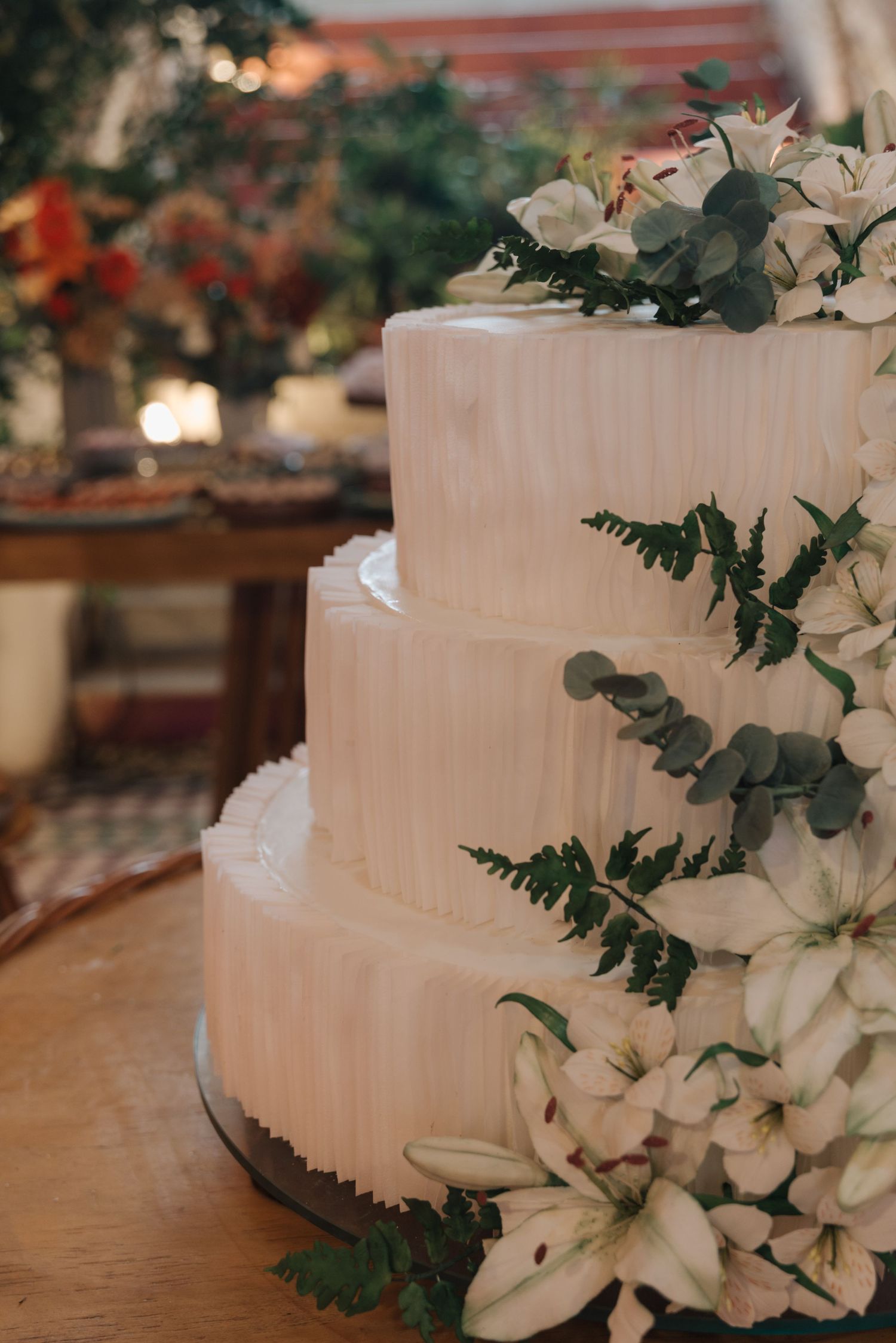 Elegant three-tiered white wedding cake decorated with lilies and greenery on a wooden table.