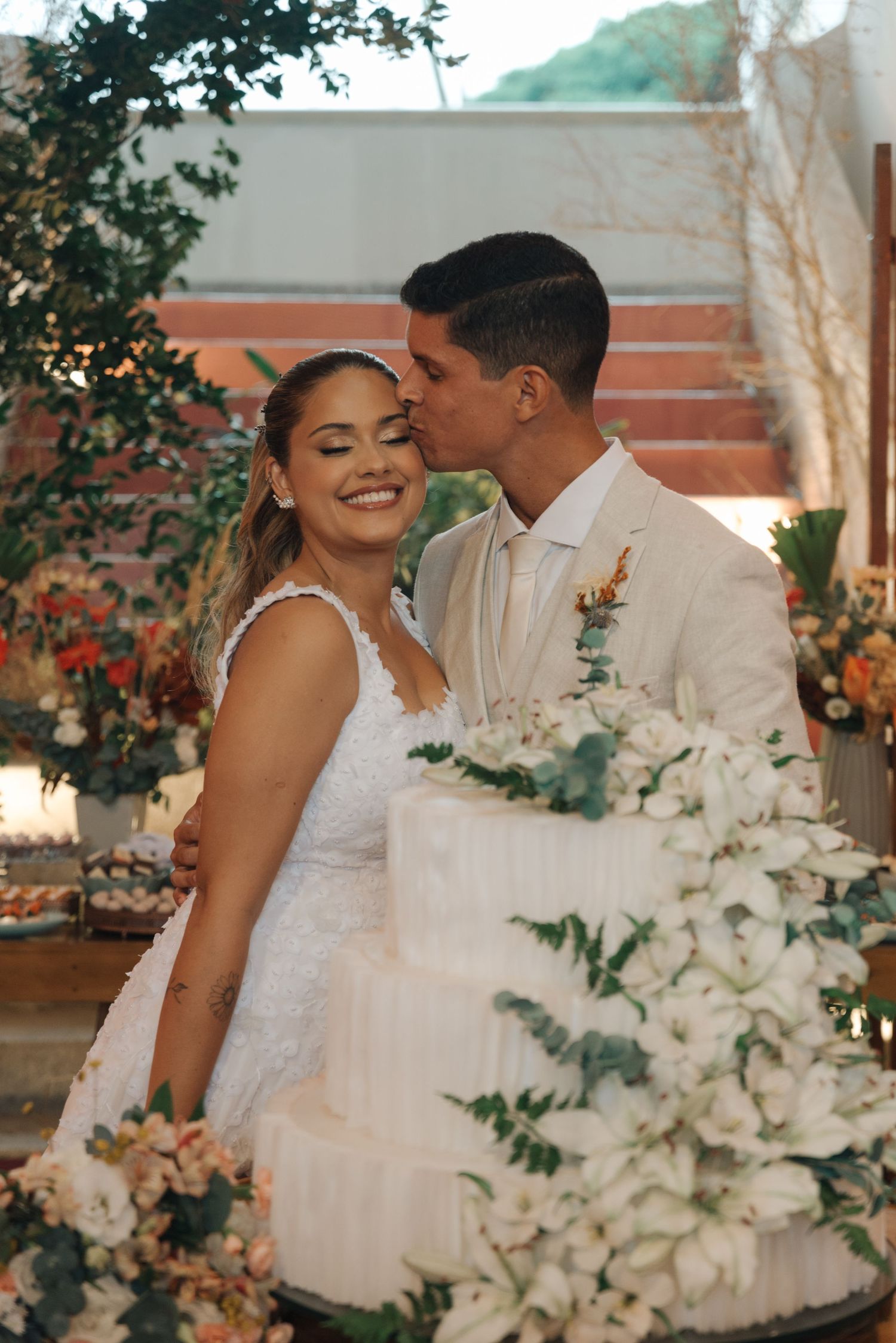 A couple shares an intimate moment next to their white floral wedding cake at an outdoor reception.