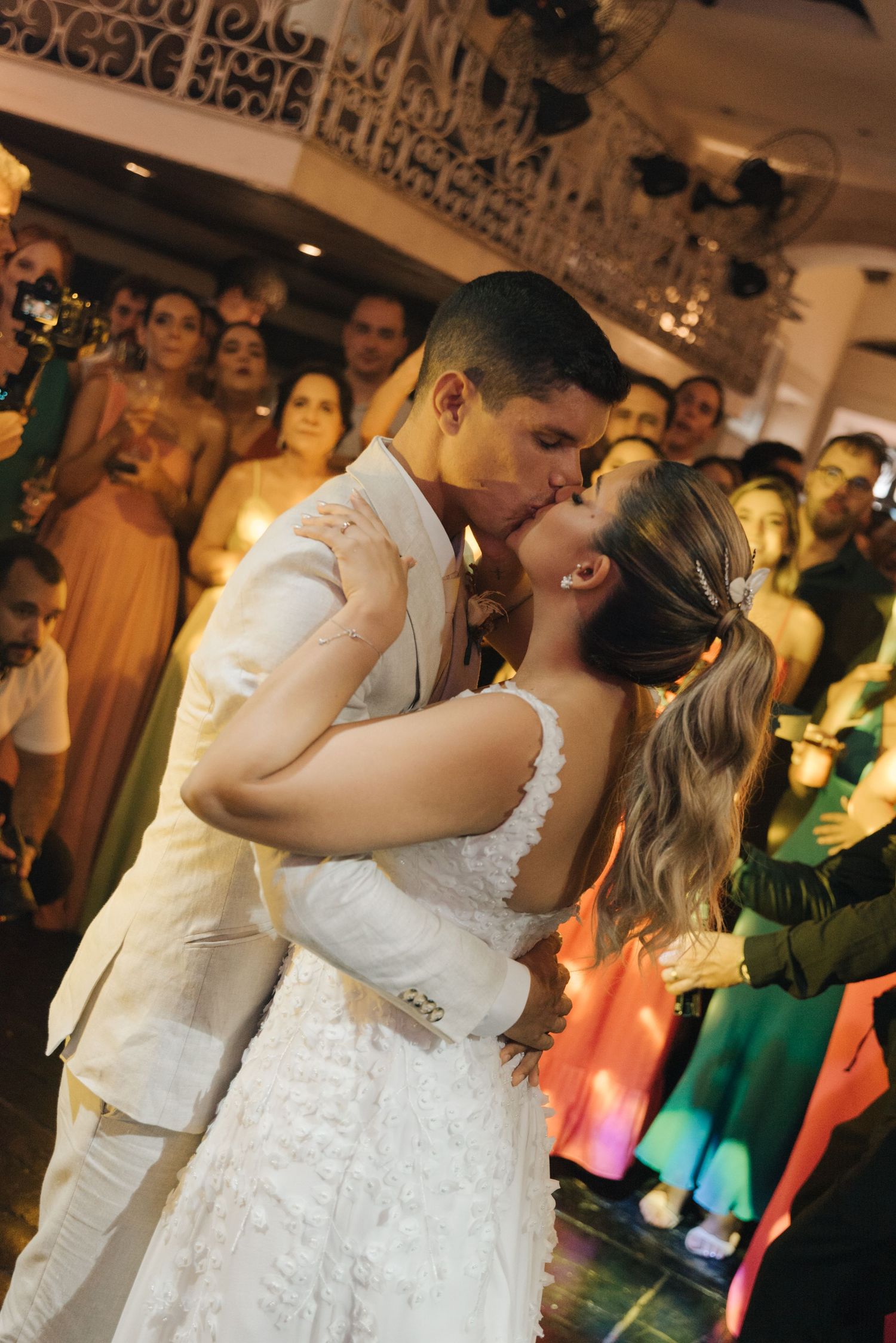 Couple shares a romantic kiss during their wedding dance surrounded by colorfully dressed guests.