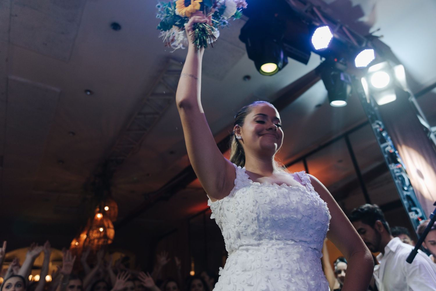 A bride in a white lace dress holds up her bouquet during a celebration at an indoor venue.