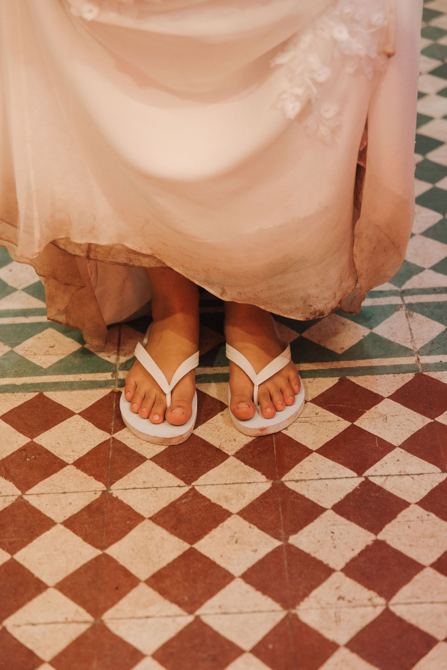 White flip flops peek out under a wedding dress on a geometric tiled floor.
