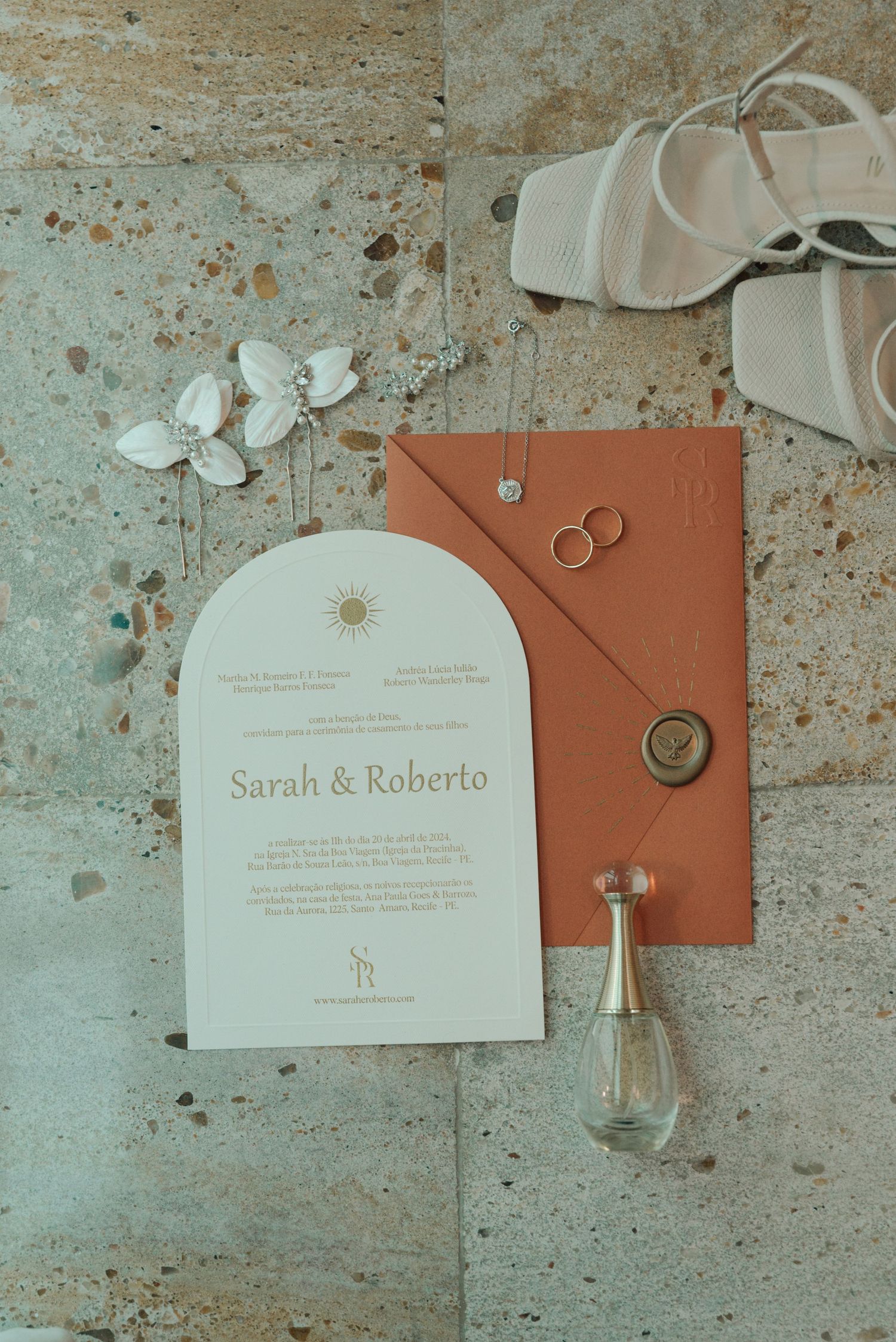 Wedding invitation for Sarah and Roberto with rings, white shoes, and perfume bottle on textured surface.