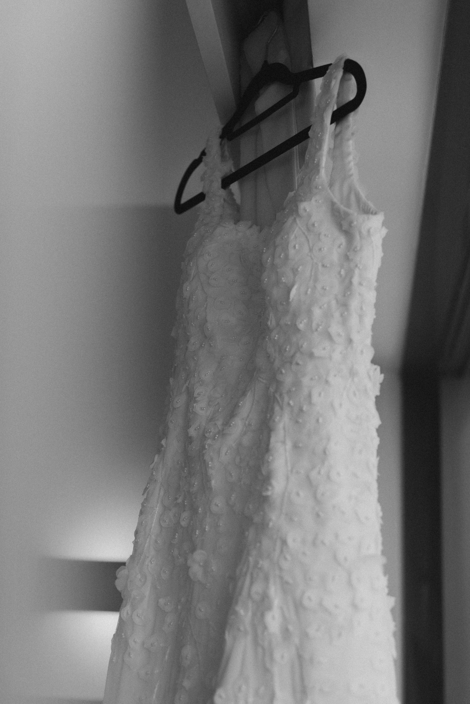 White lace wedding dress hanging on black hanger against gray wall.