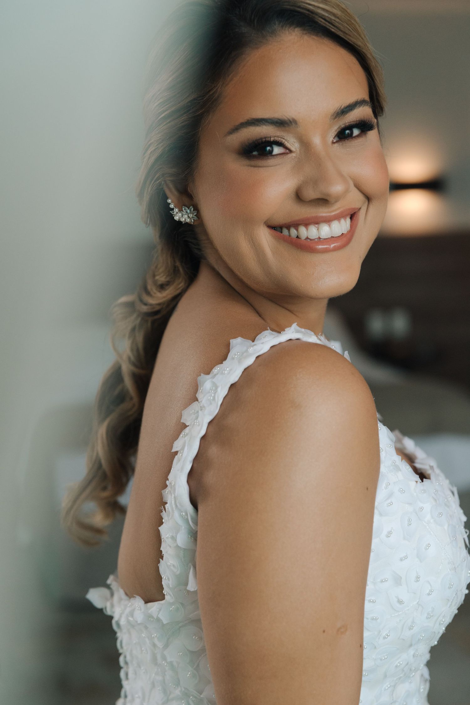 A radiant bride in a white lace wedding dress smiles warmly while showing off intricate shoulder details.