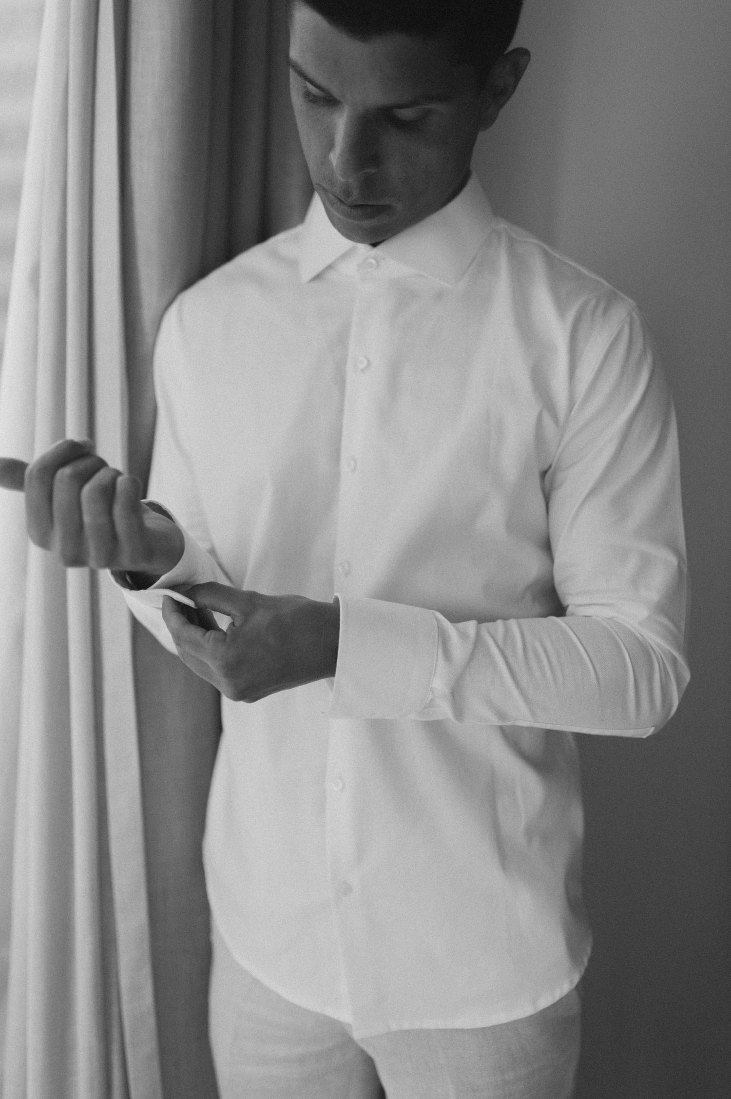 A groom in a crisp white shirt checks his phone while standing by a window.