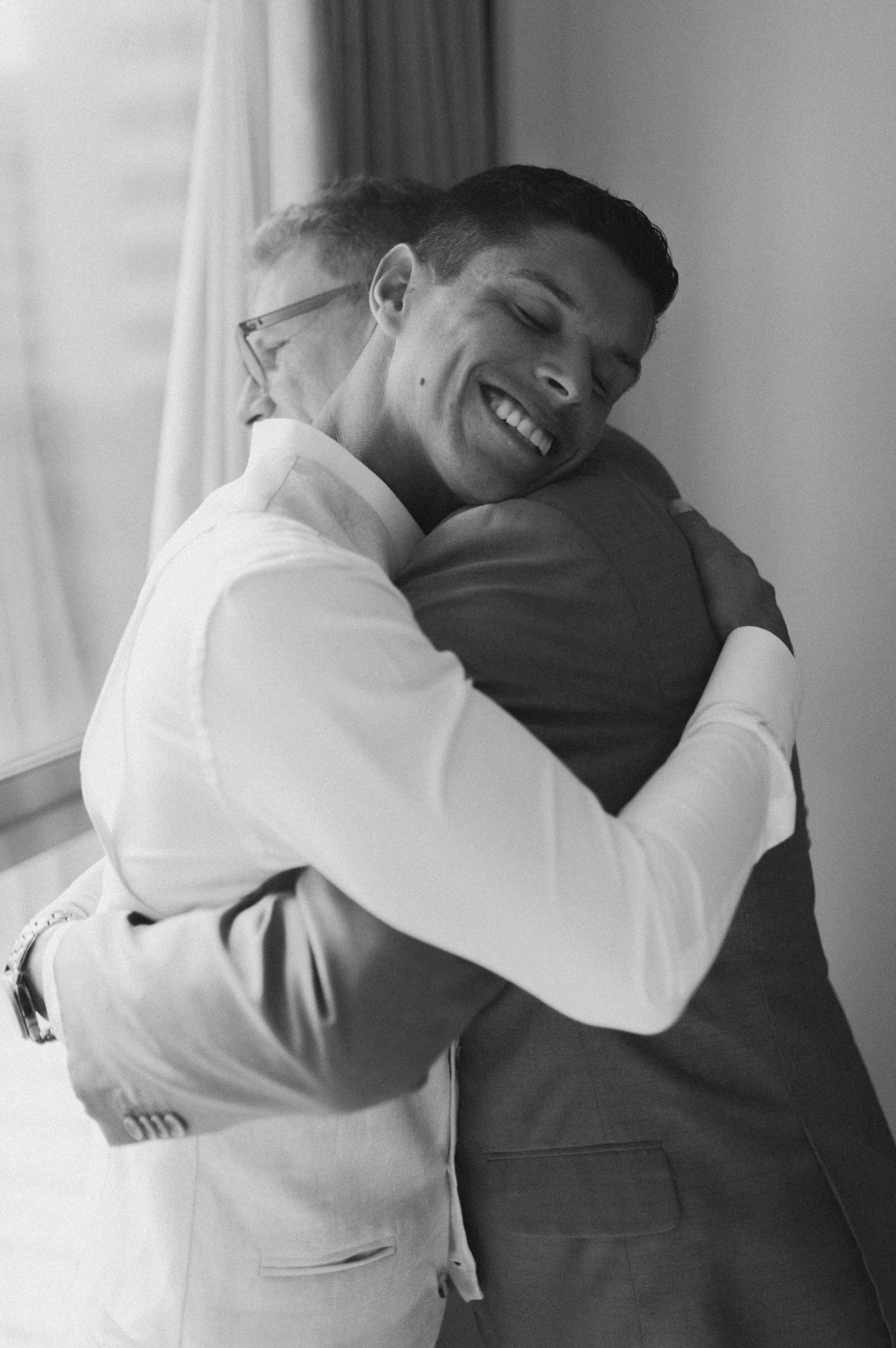 Two men in formal attire share a heartfelt embrace in black and white photography.