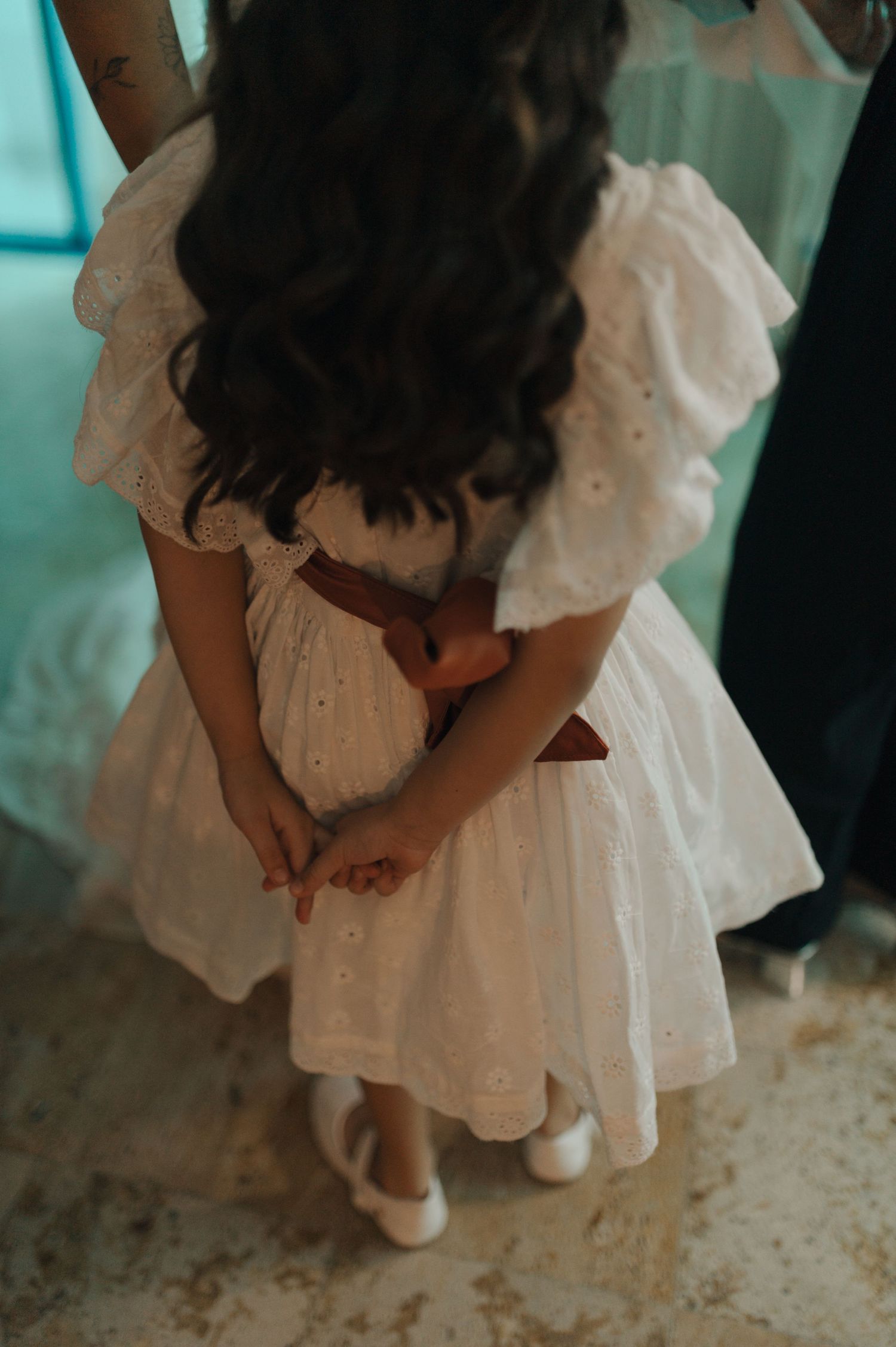 A young girl in a white dress and shoes looks down at her hands.