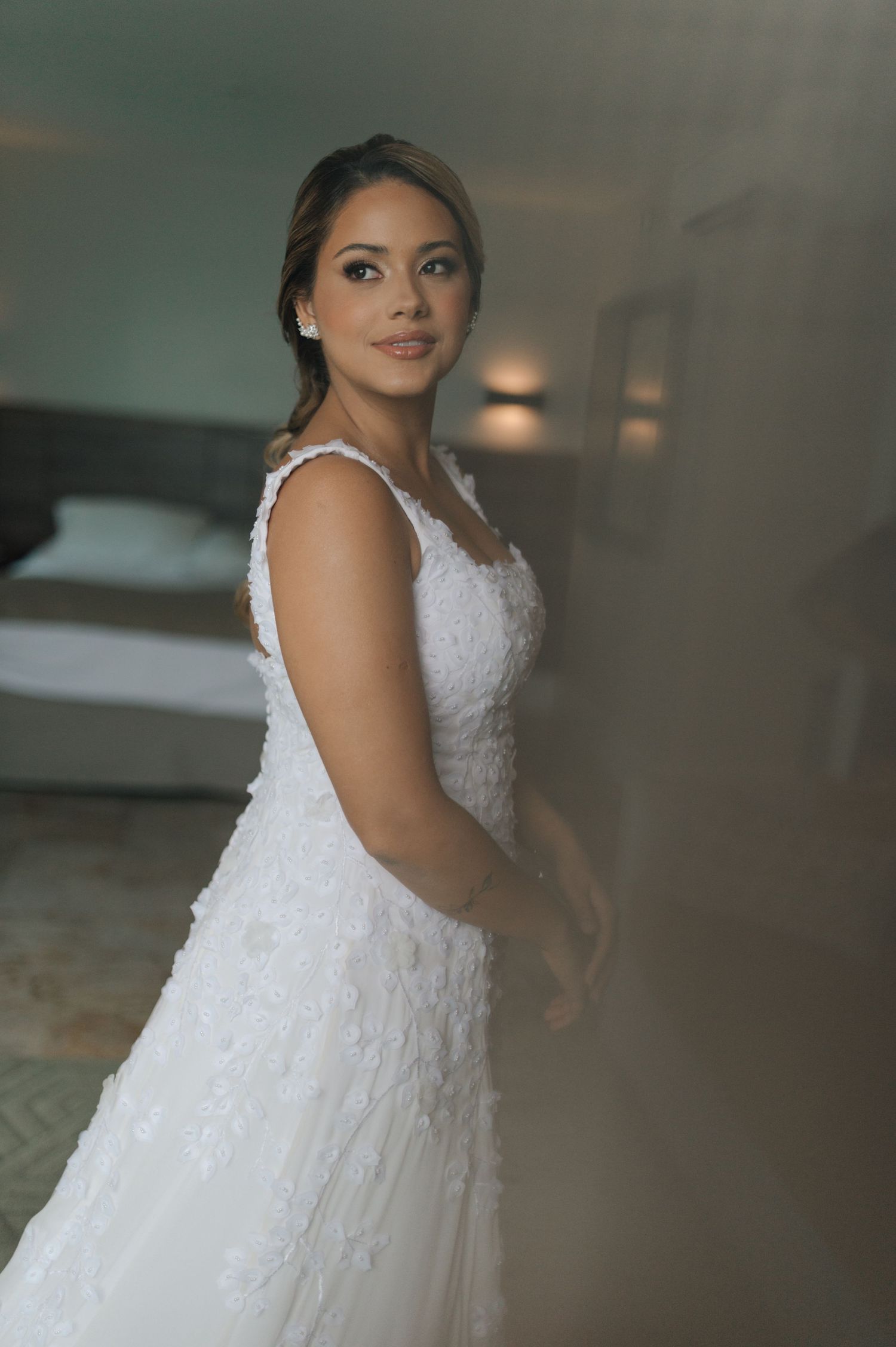 A bride in a fitted lace wedding dress poses gracefully in a softly lit room.