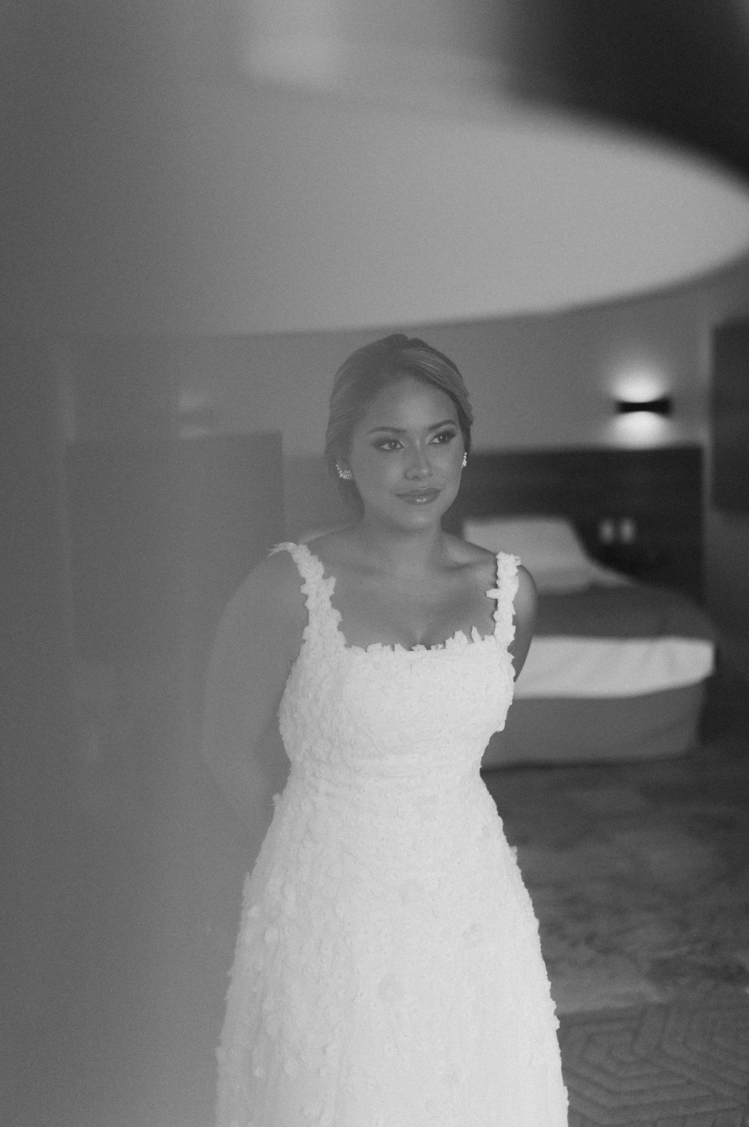 Black and white portrait of bride in lace wedding dress standing in hotel room.