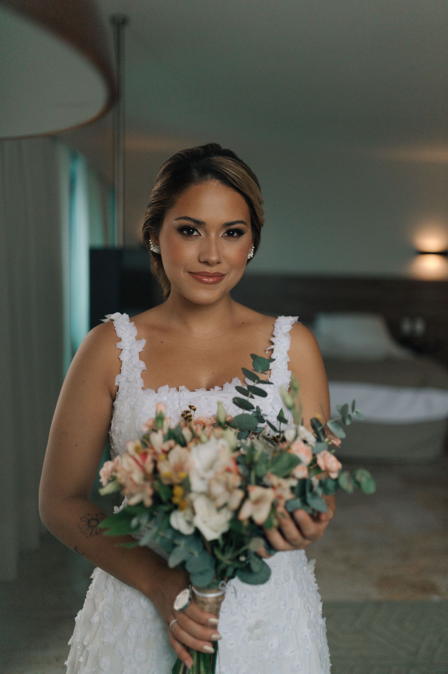 Bride holding pastel bouquet with eucalyptus in white lace gown against modern hotel backdrop.