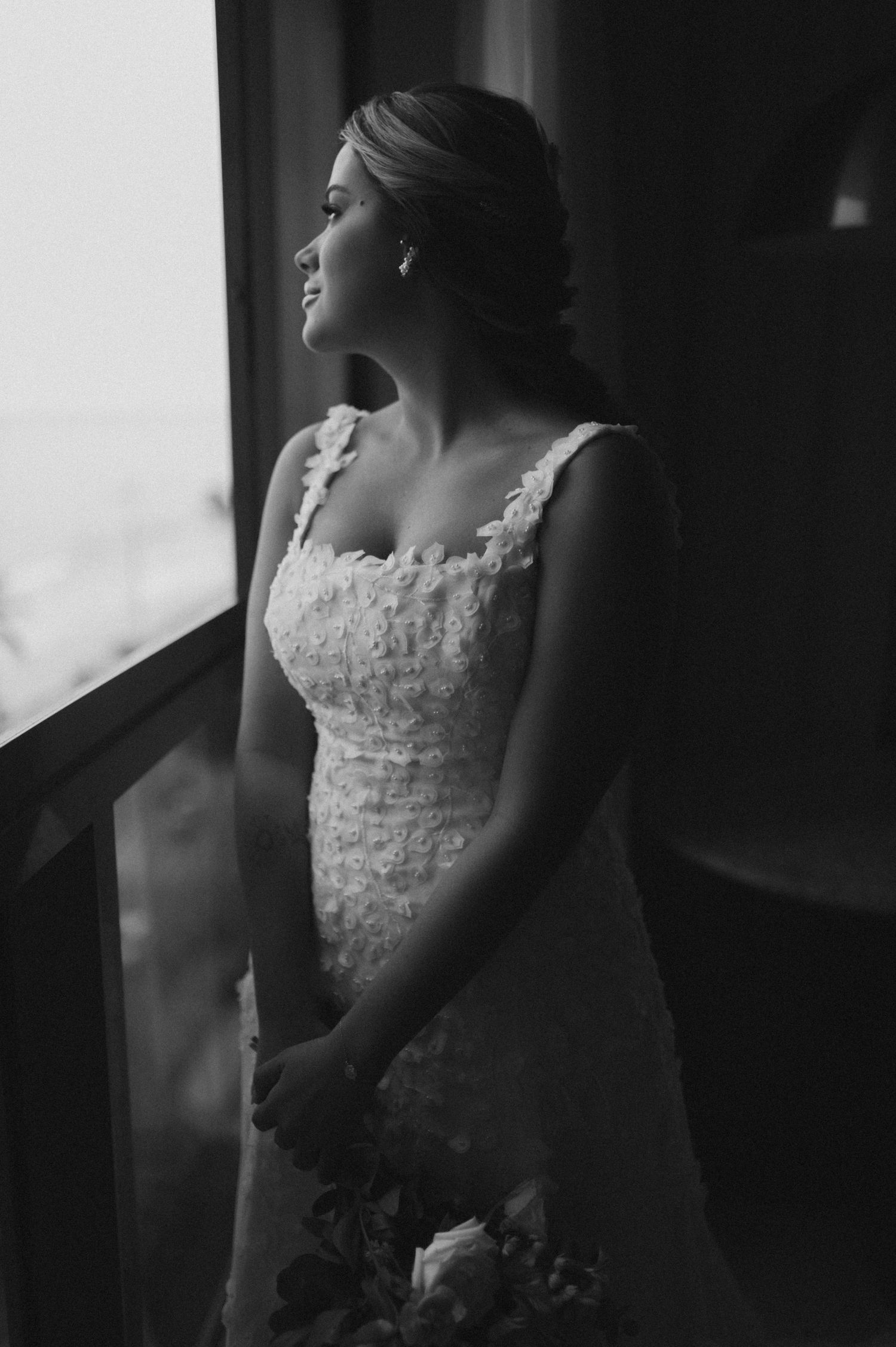 Bride in fitted lace dress gazing out window in black and white photo.