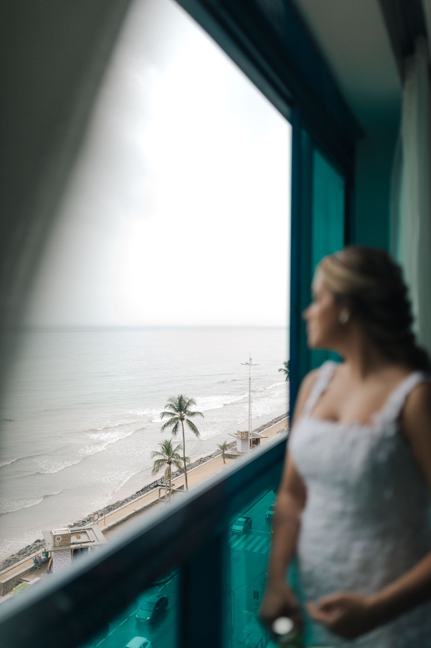Seaside bridal portrait showing bride gazing at ocean view from turquoise-tinted window.