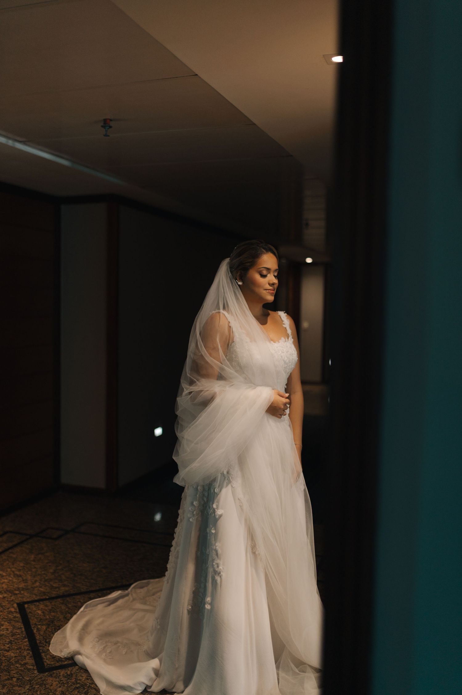 Dramatic full-length shot of bride in flowing white gown standing in dimly lit hallway.