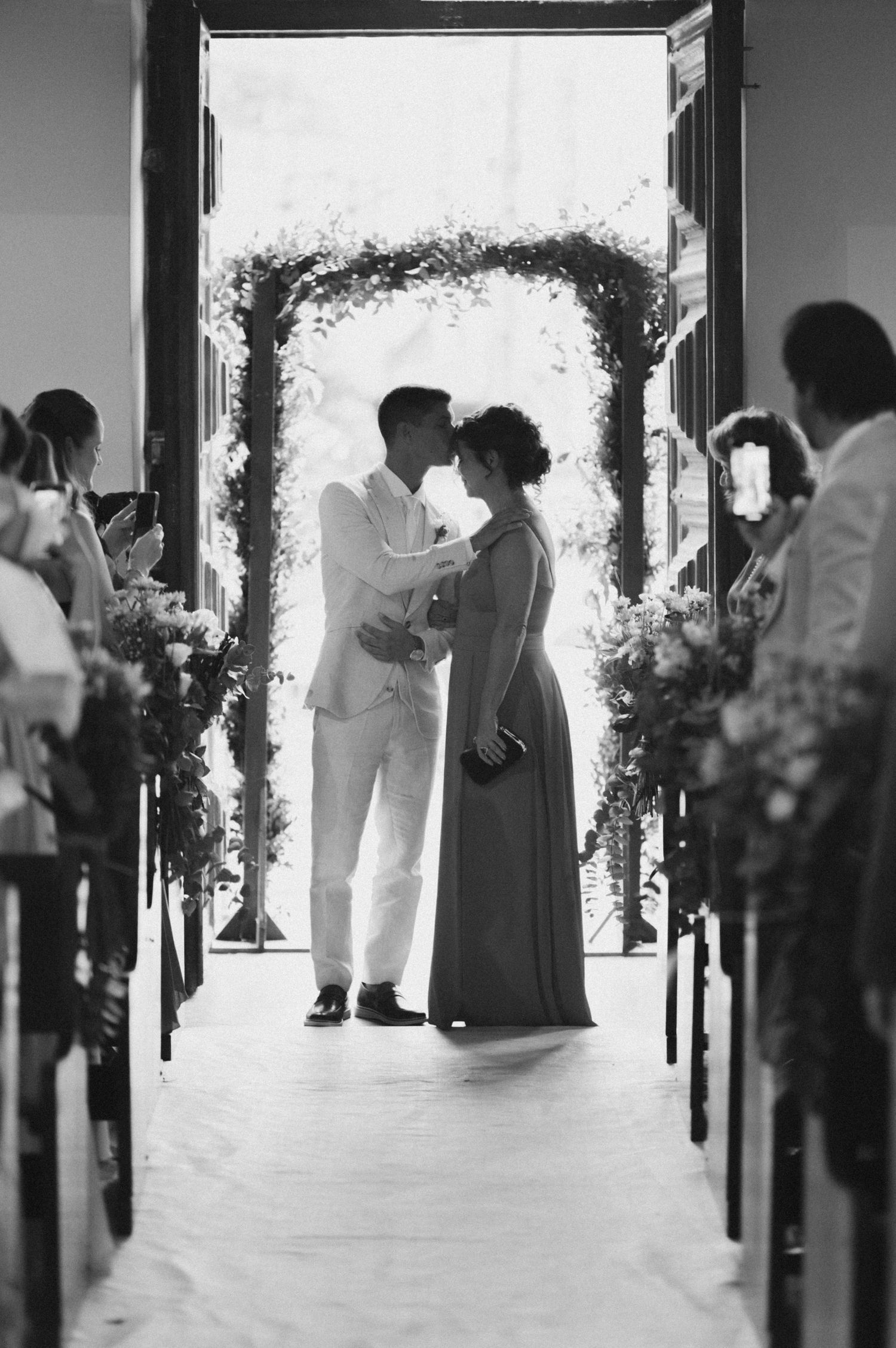 Black and white ceremony photo under floral arch with guests watching.