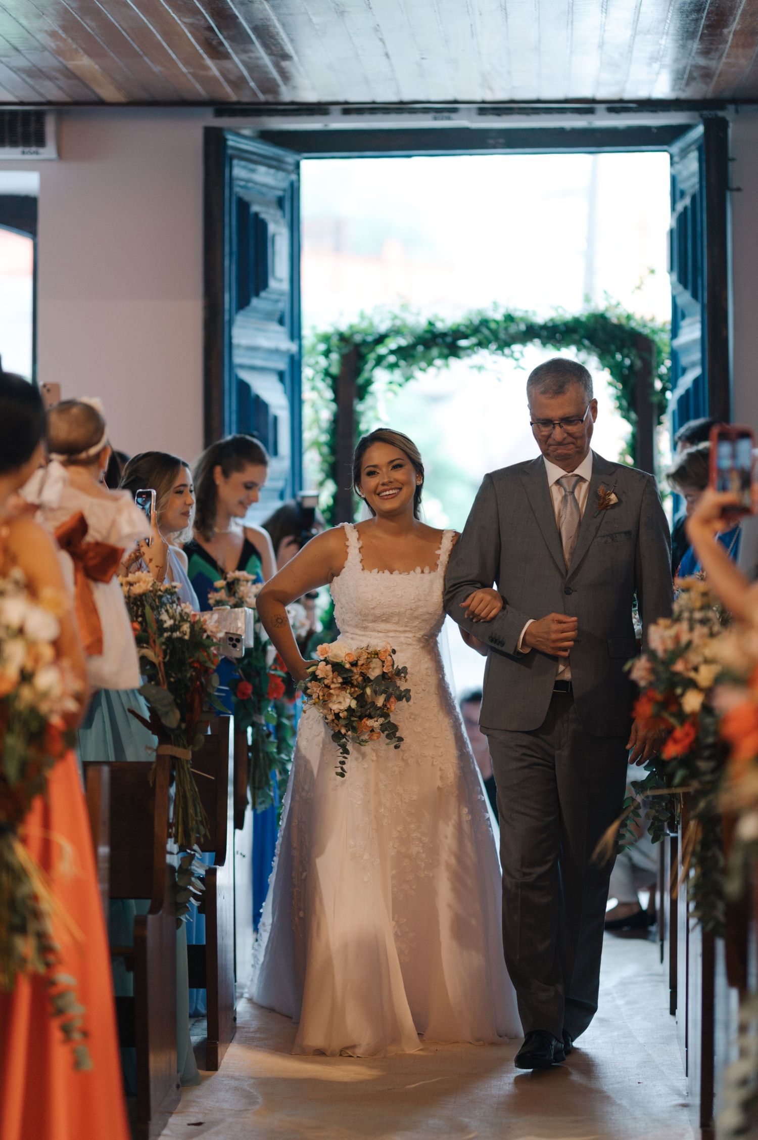 Bride walks down church aisle with escort surrounded by guests and floral decorations.