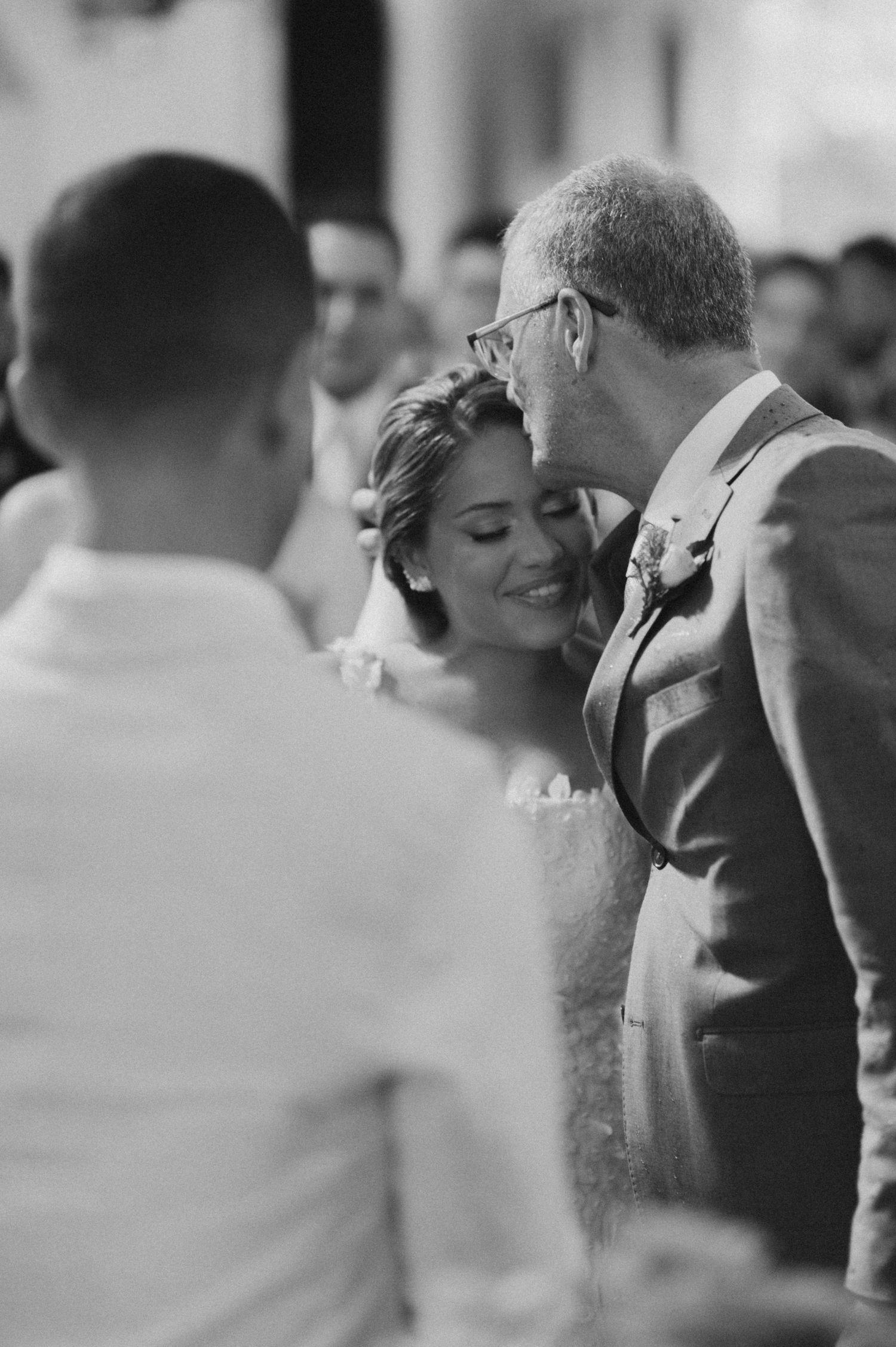 Tender moment during wedding ceremony as father kisses bride's forehead.