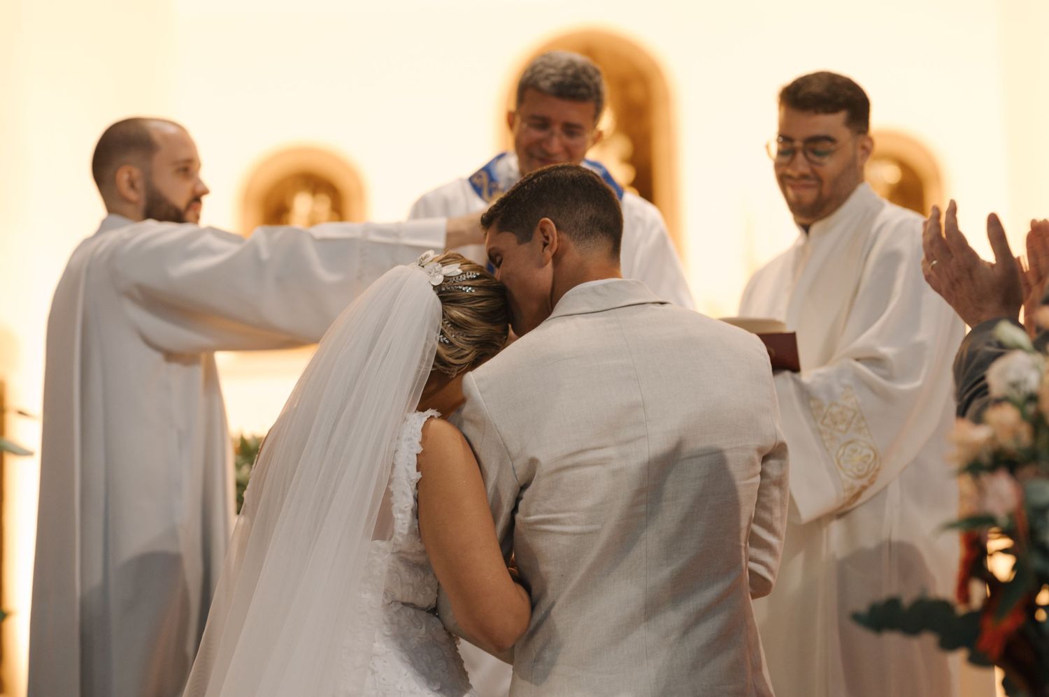 Couple embraces during wedding ceremony blessing with priests at church altar.