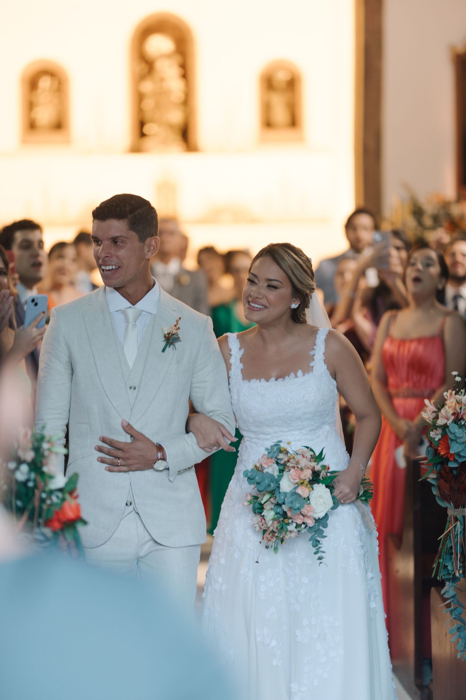 A happy wedding couple smiling while walking arm in arm with coral-colored bridesmaids in the background.