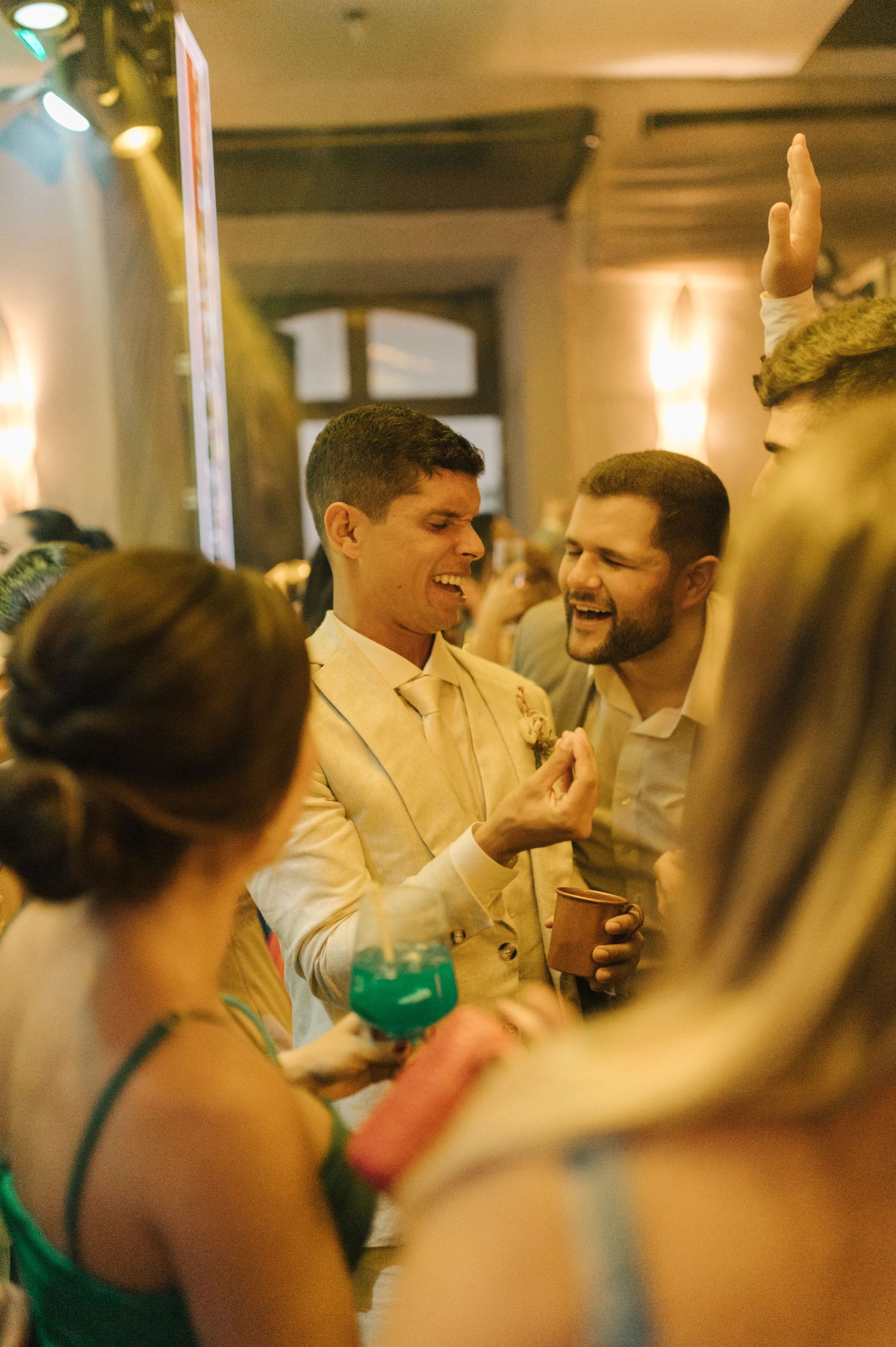 Wedding guests laughing and celebrating with drinks at an evening reception.