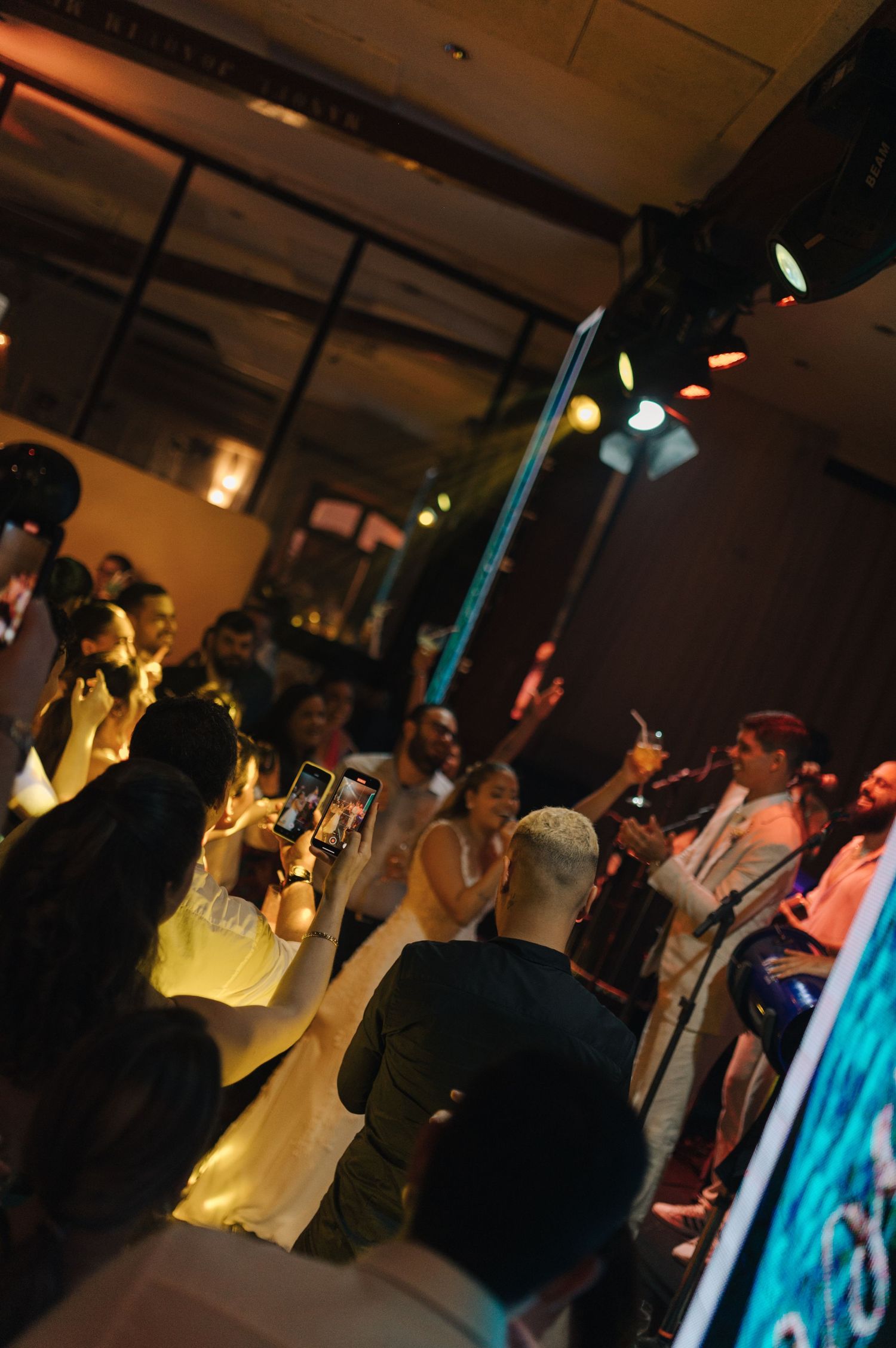 Crowd gathers on dimly lit dance floor with colorful lighting and phones capturing the moment.