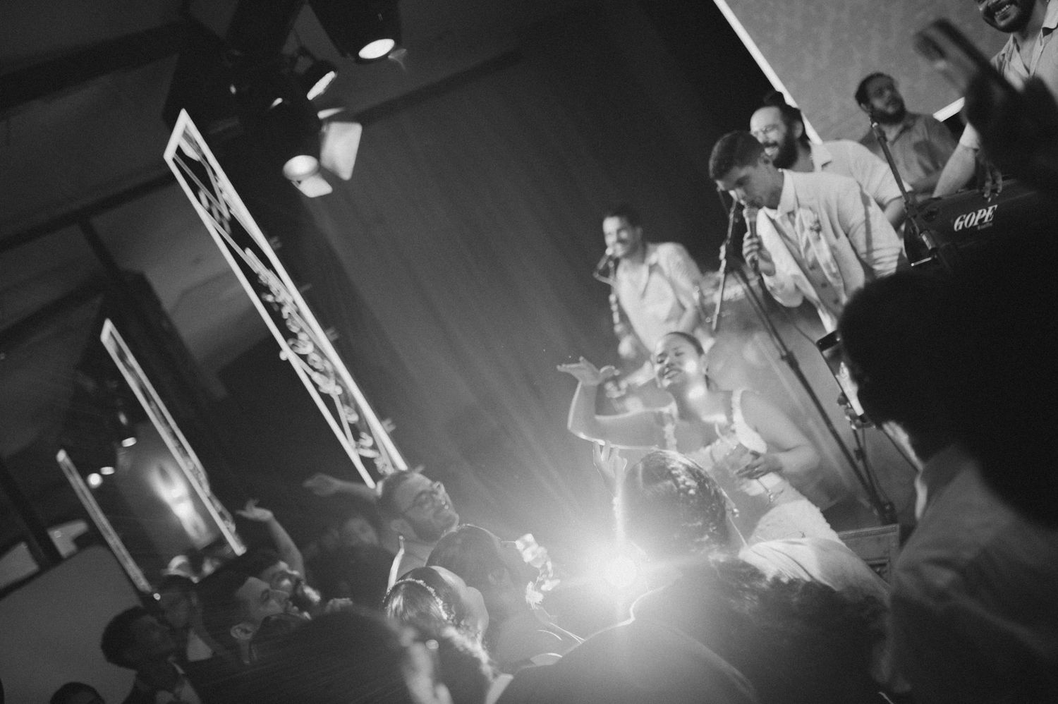 Black and white shot of performers on stage with dramatic spotlight and audience silhouettes.