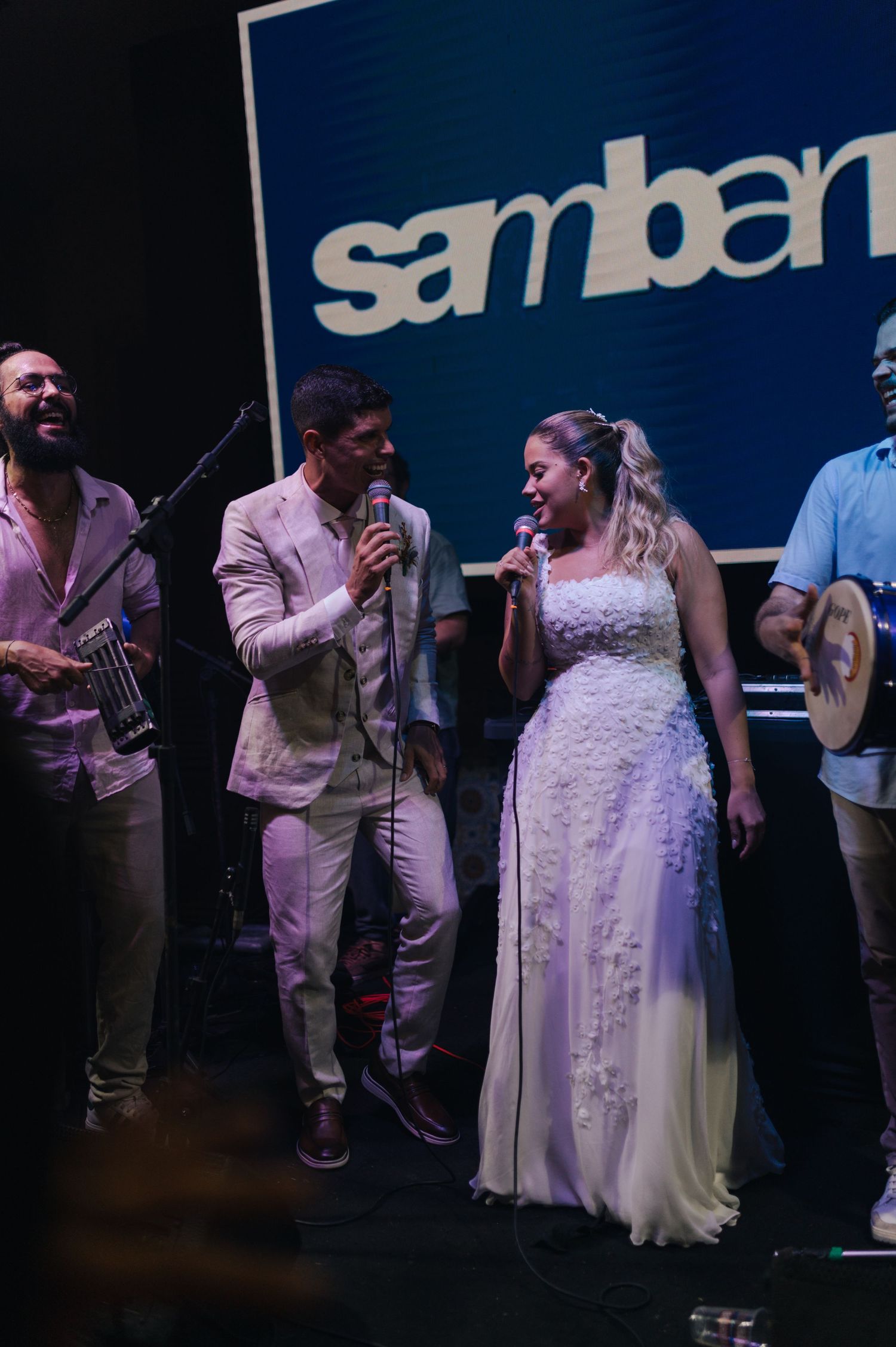 Band performs on stage with Samban logo backdrop while wearing light colored outfits.