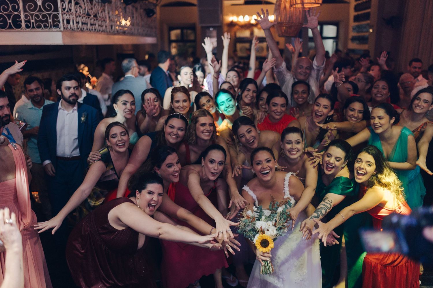 Large group of wedding guests posing together and reaching toward the center for a celebratory photo.