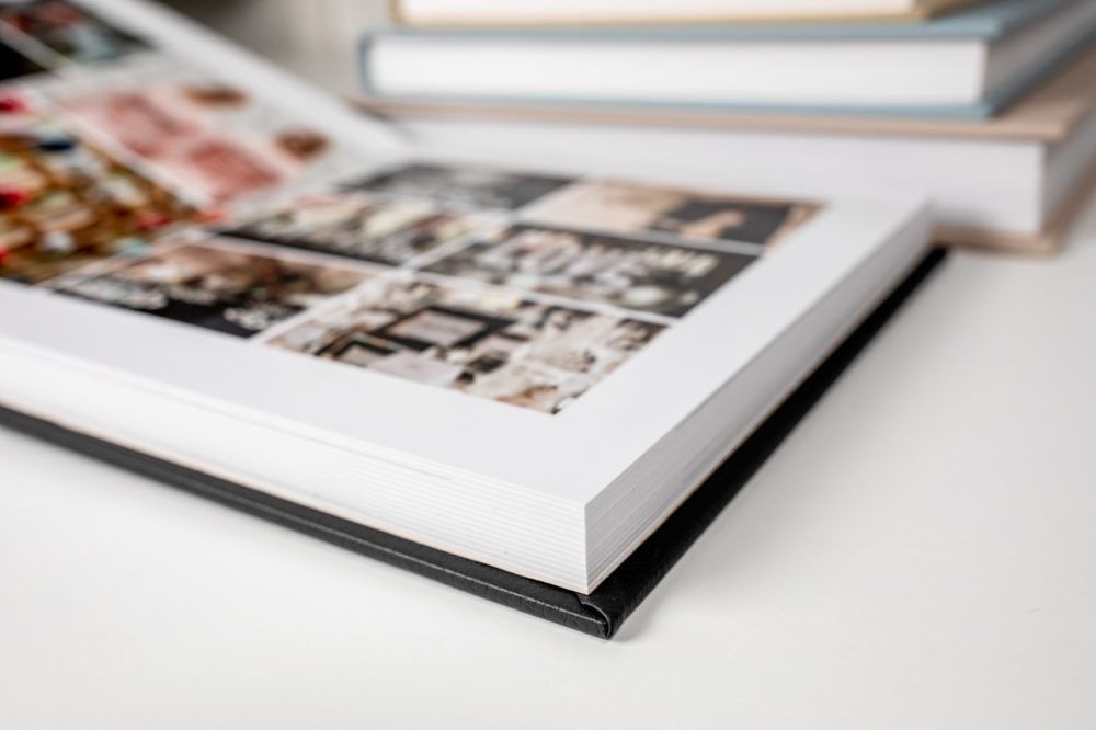 White hardcover photo book with a corner shown on a light surface, displaying interior pages with multiple images.