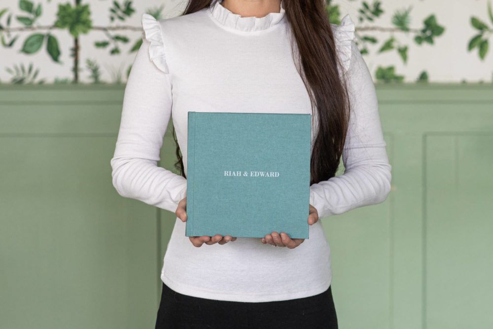 Someone in a white sweater holds a teal-colored hardcover photo album against a sage green paneled wall with leaf decor.