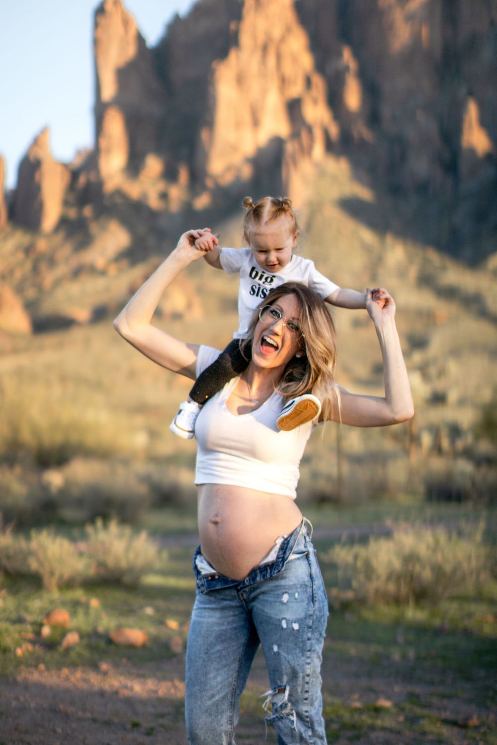 An expectant mother smiles big while holding her toddler on her shoulders.