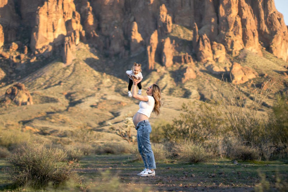 An expectant mother lifts her toddler into the air, standing before the Superstition Mountains.