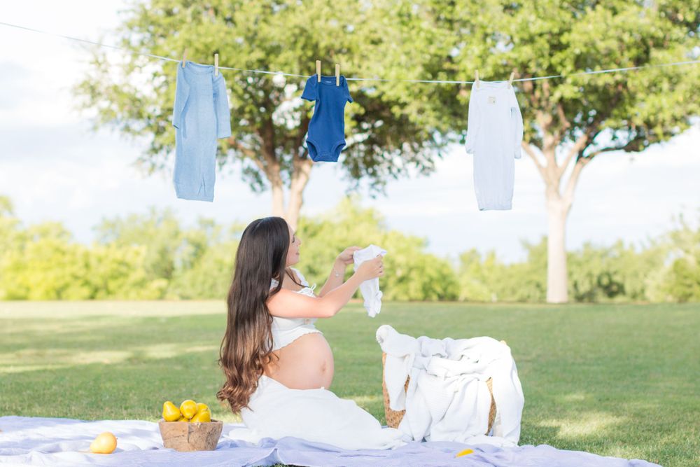 An expectant mother holds up a baby onesie, deciding where to hang it on the clothesline behind her.