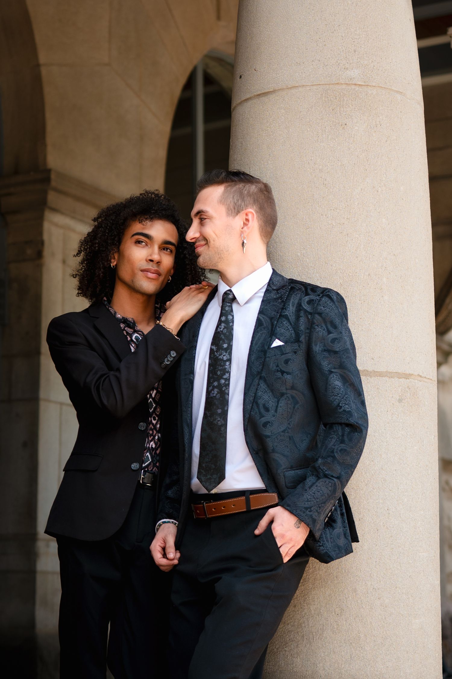 Two people in formal attire share an intimate moment against stone pillars in an elegant architectural setting.