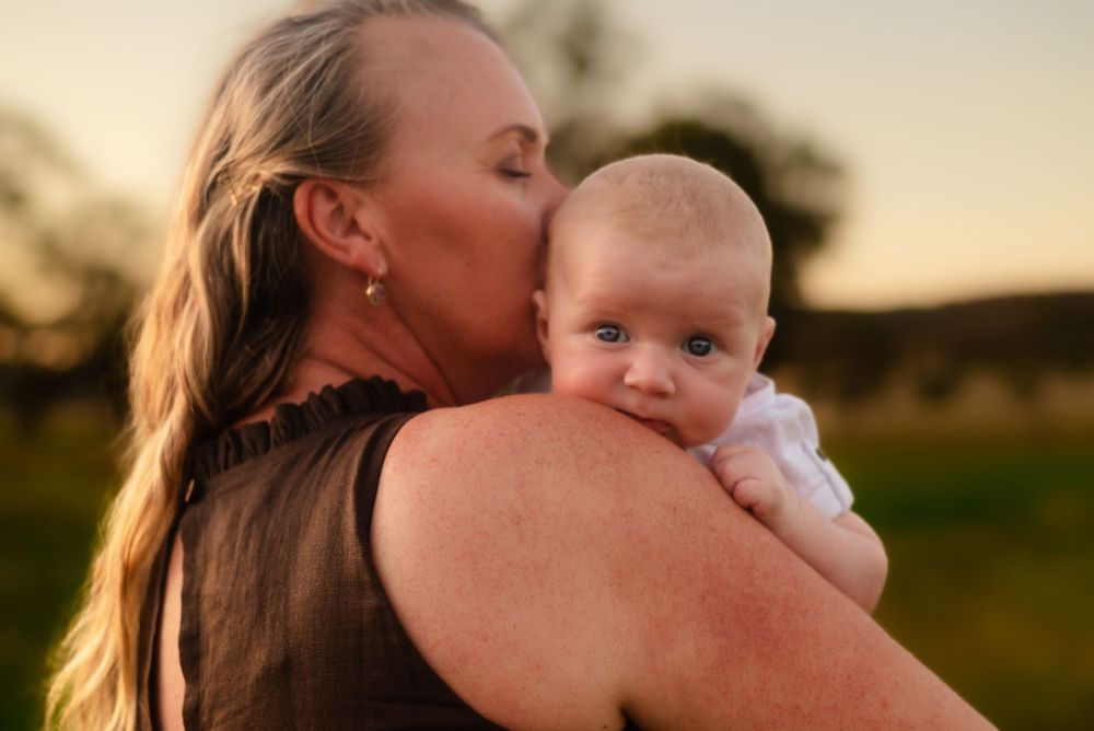 Parent holding and kissing baby during sunset outdoors.