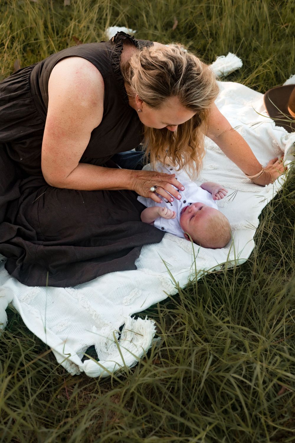 Parent playing with baby on white blanket in grassy field.