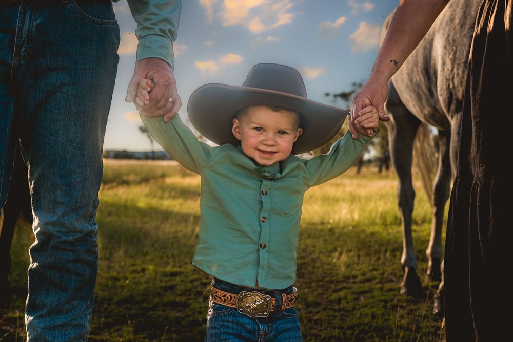 Child in cowboy hat holds hands with adults beside horse in sunset field.