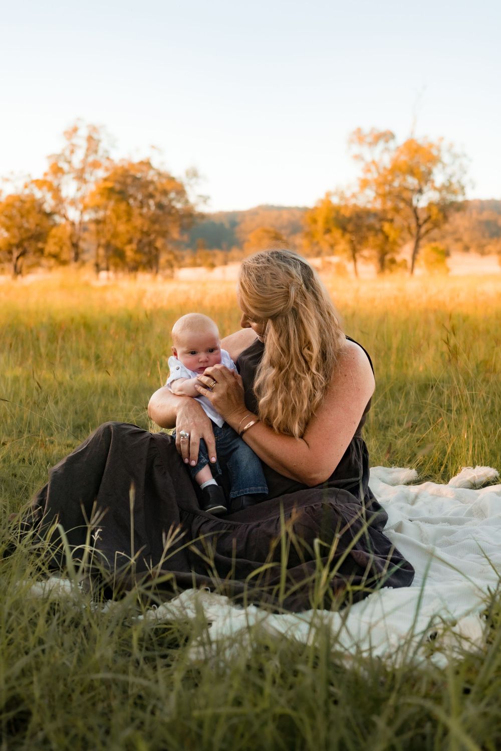 Mother holding baby sits on white blanket in golden field at sunset.