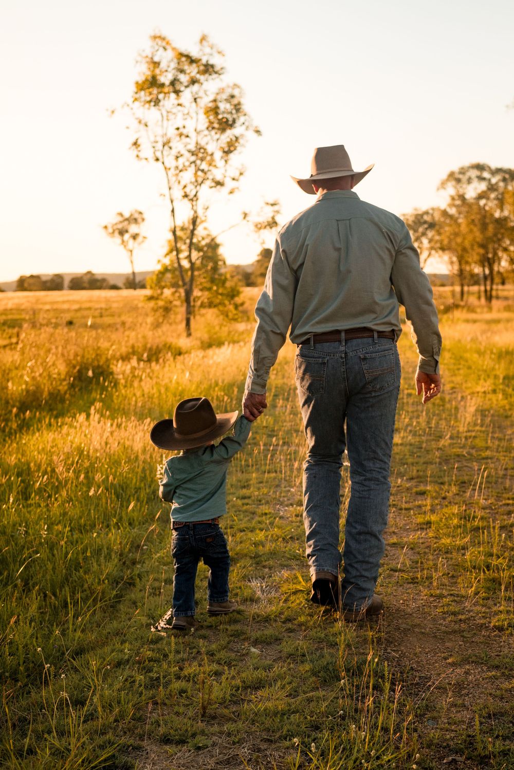 Person in cowboy hat walking with small child on country path at sunset.