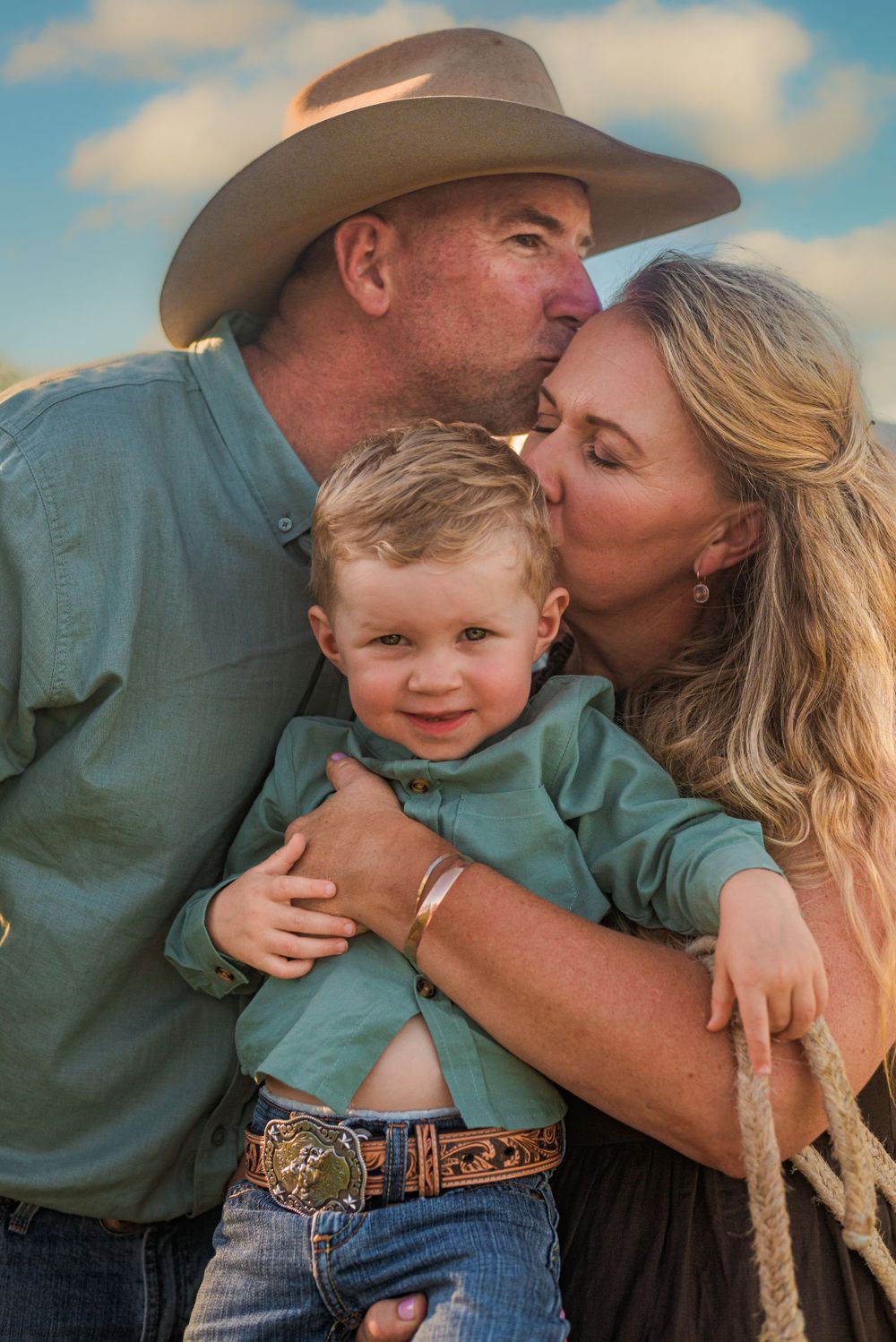 Parents kiss their smiling toddler in cowboy-themed family portrait at sunset.
