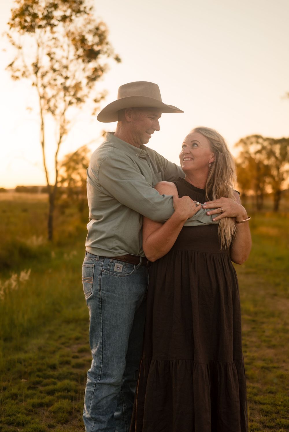Couple in western attire embracing during golden hour sunset on a rural farm.